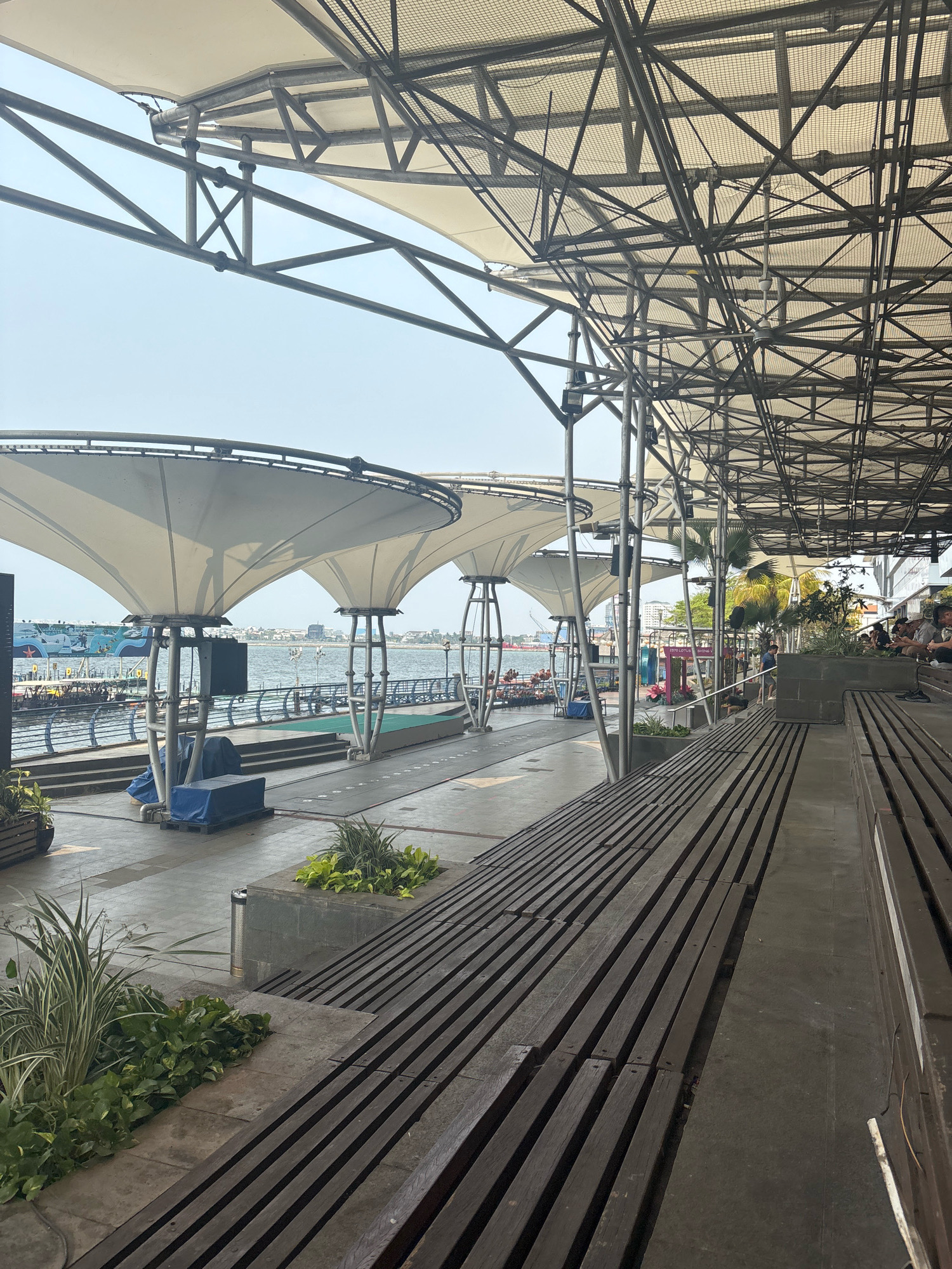 The image depicts an outdoor seating area overlooking a waterfront in North Jakarta, Indonesia. The seating consists of long, dark wooden benches arranged in tiered rows, suggesting a public space designed for viewing or events. Above the seating, a large, modern canopy structure is supported by a complex network of metal beams and poles, providing shade. The canopy itself is made of white fabric.
In the background, the waterfront is visible, with calm water and several boats docked or moving. A promenade runs along the water's edge, lined with railings and some greenery. Beyond the waterfront, a cityscape with buildings and palm trees is visible under a bright, hazy sky.
Several people are present in the scene. Some are seated on the wooden benches in the foreground, appearing to be relaxed and casually dressed. Others are visible further back, near the waterfront, suggesting various activities such as strolling or gathering. A few large speakers or sound equipment are placed on the ground near the seating area, possibly indicating preparations for an event. Blue tarps cover some equipment or structures near the waterfront. The overall atmosphere is one of a bustling urban waterfront area, likely a recreational or entertainment destination.