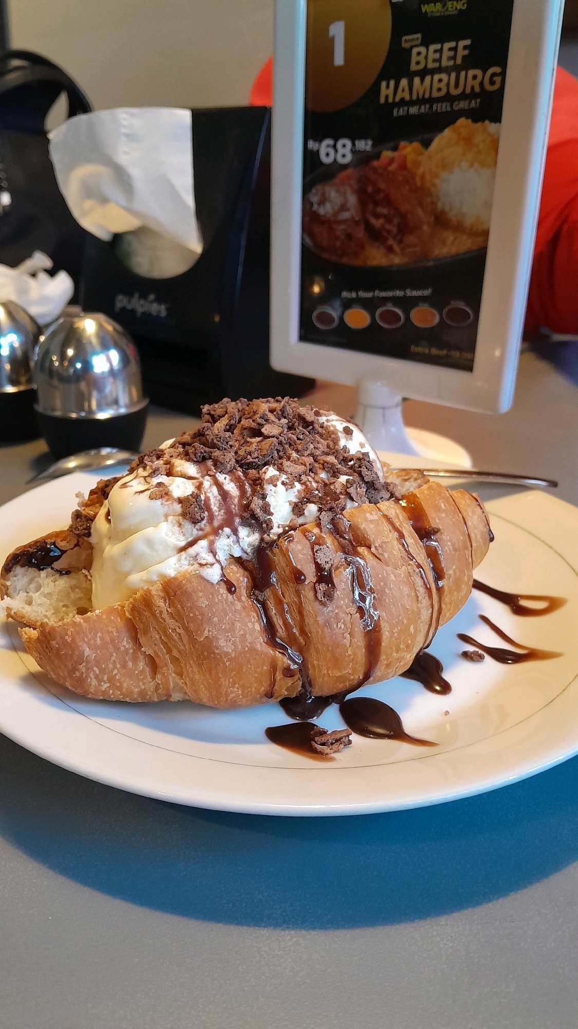 A close-up, eye-level shot captures a dessert item resembling a croissant filled with vanilla ice cream and topped with chocolate shavings and syrup, resting on a white plate. The plate sits on a blue-grey surface. In the background, a digital menu board displays information about a "Beef Hamburg," featuring a price of "Rp 68,182" and images of food. To the left of the menu board is a tissue dispenser with the logo "pulpies." The environment appears to be an indoor dining area. The image does not provide enough visual information to ascertain a specific location in Bandung City, Indonesia.