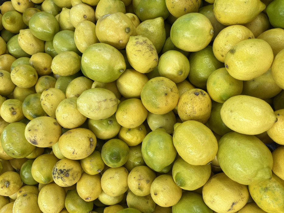 A dense collection of lemons fills the frame, showcasing a range of yellow and green hues. The lemons vary in size and shape, with some displaying slight imperfections or discoloration. The pile is tightly packed, creating a textured surface of citrus fruit. The lighting is bright and even, highlighting the natural colors and textures of the lemons.