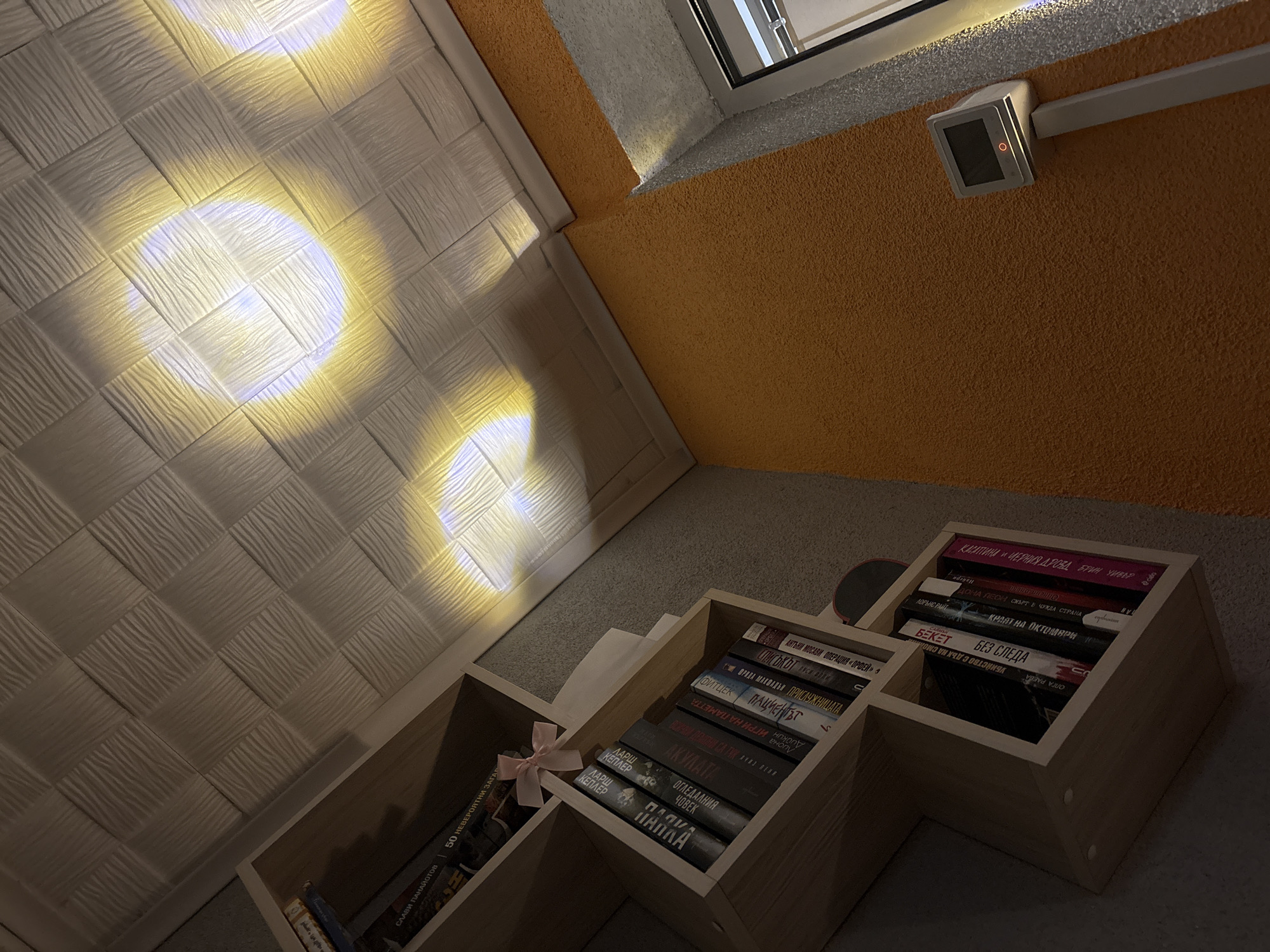 The image shows a set of wooden shelves containing books, arranged in an interior space with textured walls and a glimpse of an exterior environment through a window. The books are various titles in Bulgarian and English, including works by authors such as Slavi Panayotov, Anthony Mosawi, and Fitzek. A light source creates bright spots on the textured wall, which gives the scene a unique lighting quality. The wooden shelves are positioned at an angle, emphasizing the oblique perspective of the shot. The overall setting has the feel of a reading nook, possibly in a home or small library. I do not know where this image was taken or the city in which it exists.
