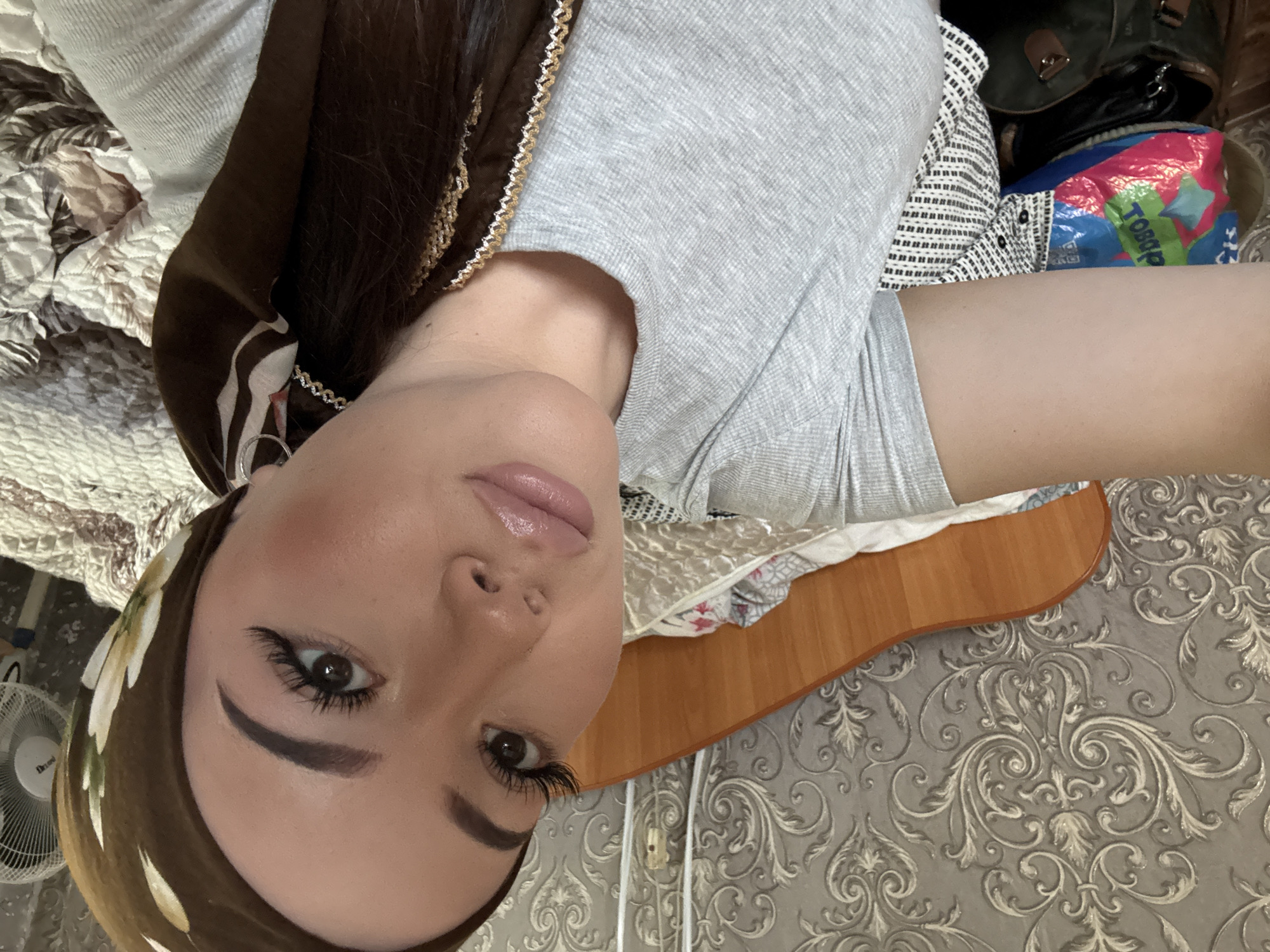 A woman with fair skin, brown eyebrows, and dark eyelashes is shown wearing a brown headscarf with a floral pattern and a light gray t-shirt. She is in an interior location, possibly Коноваловка, Russia, given the presence of specific items. The background features bedding with a patterned comforter, a patterned wall, a wooden surface, a white fan in the lower left corner, and a bag with red and blue colors.