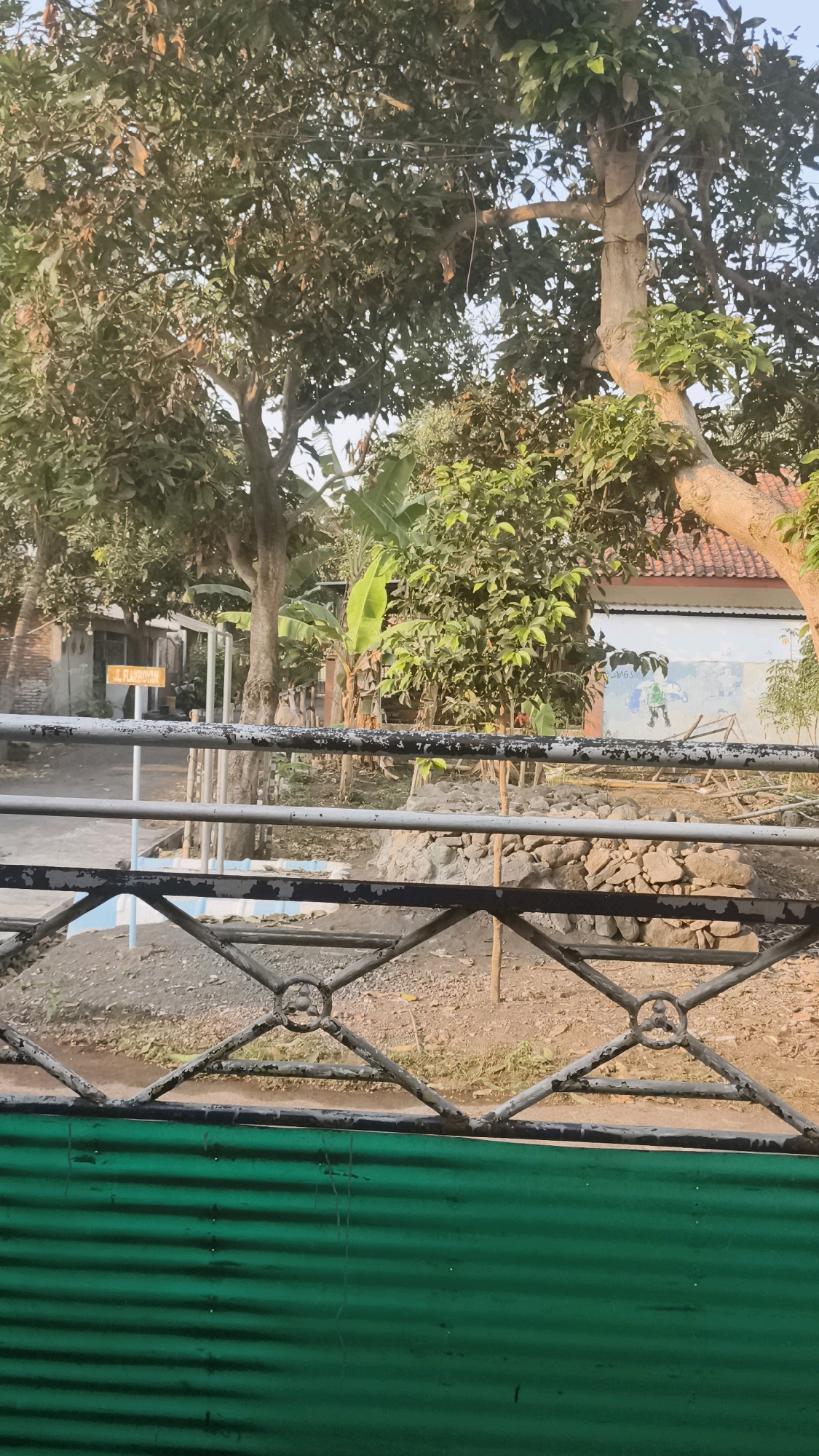 The image shows a view through a metal gate and green corrugated metal barrier of a yard in Kutorenon, Indonesia. In the background, a building with a red tile roof and a white wall adorned with a mural of figures is visible. The yard contains trees, including a large tree with a thick trunk and branches, and banana plants. A sign with Indonesian text, "JL. RAYAHOYO," is posted on a pole. Rocks are piled up near the building. The ground is covered with dirt and gravel. The sky is clear and blue.