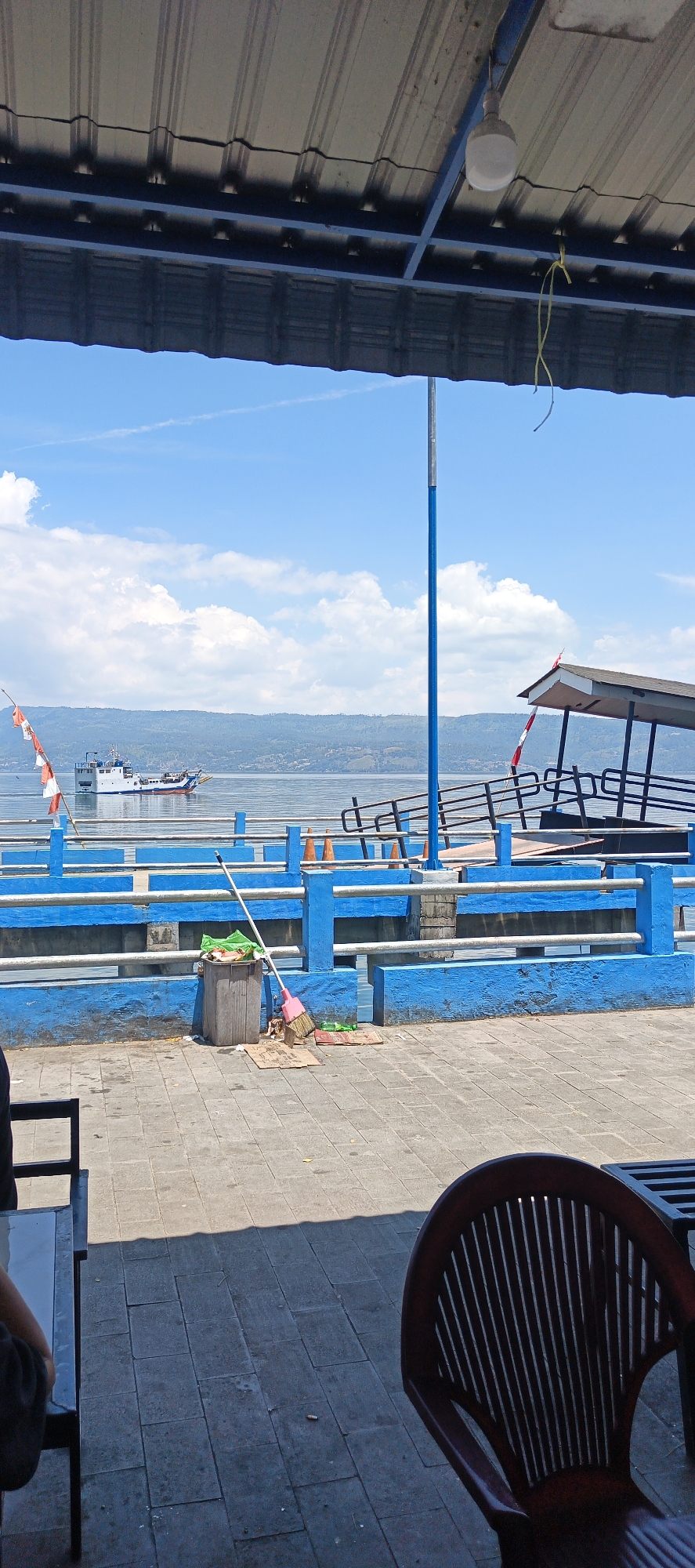 The image shows a waterfront scene with a blue railing and a boat in the distance. The scene appears to be located in Tigaras, Indonesia, given the flags visible. In the foreground, a broom and dustpan are leaning against a trash can. A dark wooden chair is positioned at a table in the lower right corner of the image, casting a shadow. The sky is blue with scattered clouds.