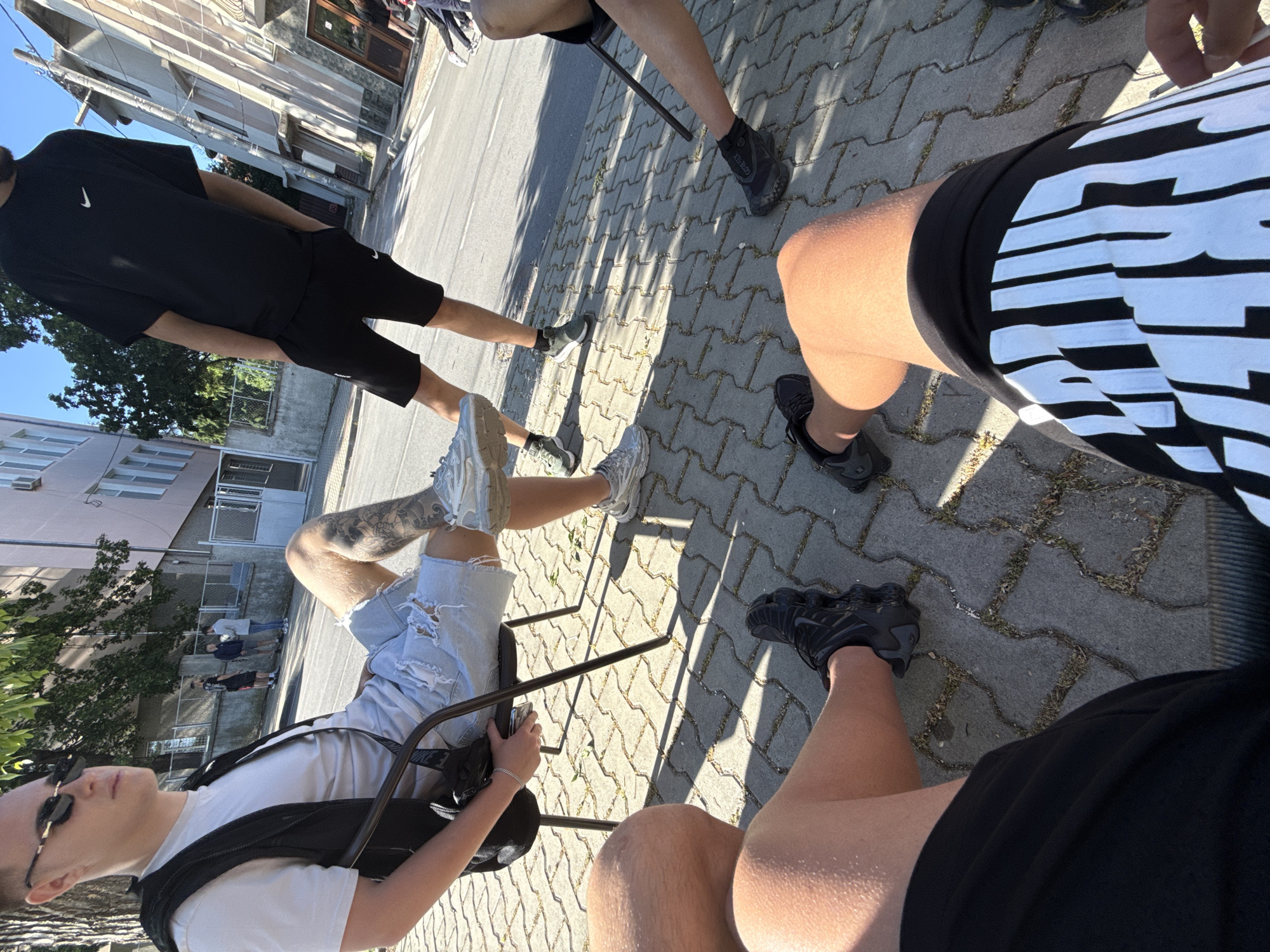 Four people are gathered outdoors, likely on a paved area in Elhovo, Bulgaria. One person wears sunglasses, a white t-shirt, ripped denim shorts, and has a tattoo on their leg. Another person wears a black Nike t-shirt and shorts. A third individual is wearing shorts with white text. The fourth individual has dark shoes. All four are seated. The surrounding environment includes buildings, trees, and a street.