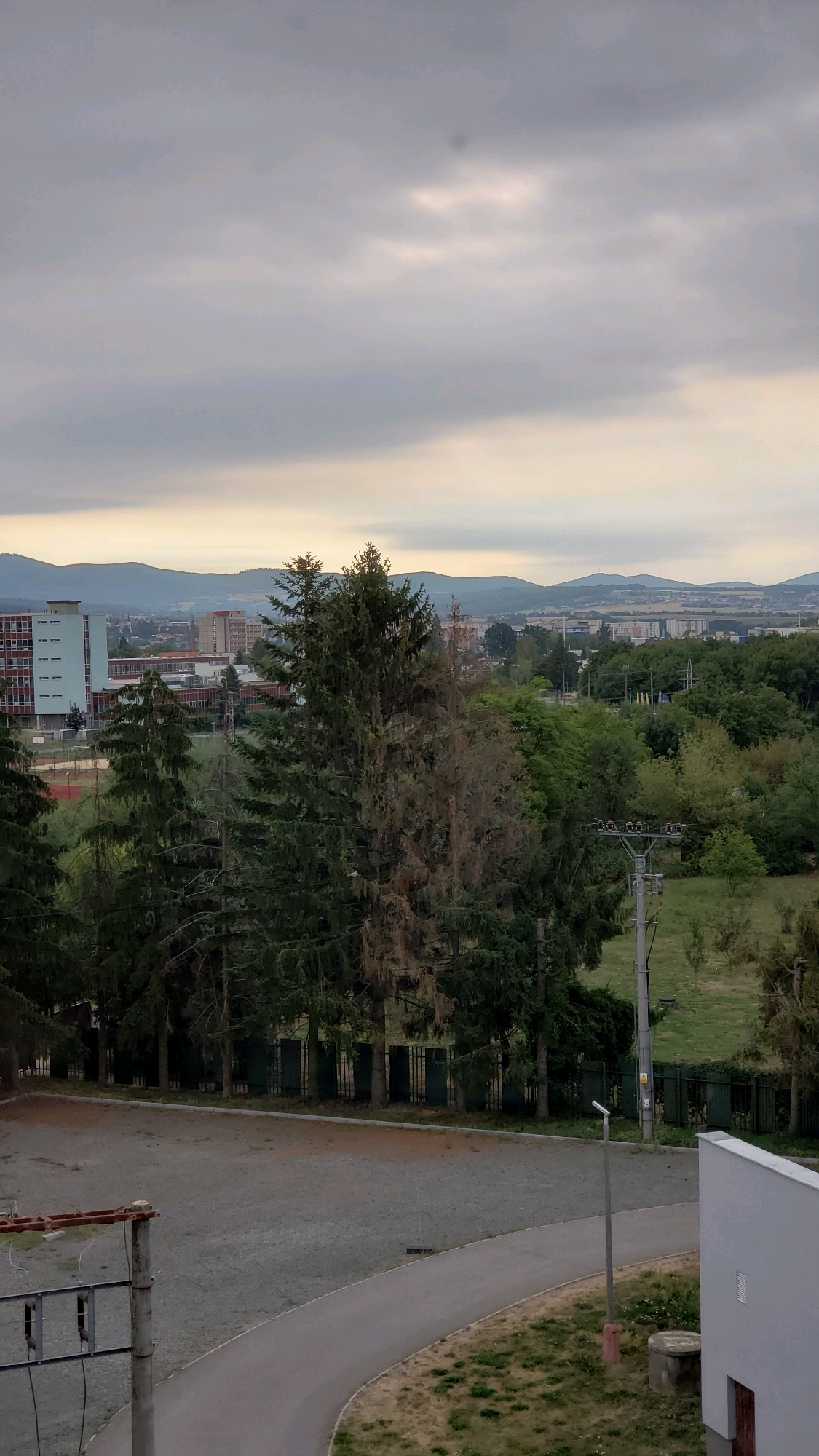 The image captures a view from a slightly elevated position, looking out over an urban landscape. In the foreground, a gray, gravel-covered area is bordered by a paved pathway that curves to the right. A metal pole with electrical equipment is visible on the left side of the pathway, and another pole with a street light stands further down. A white building with a vent is partially visible on the right.
Beyond the immediate foreground, a line of tall evergreen trees, some appearing darker green and others lighter or even brown and dry, dominates the middle ground. Behind the trees, various buildings can be seen, including a multi-story, light blue and red brick building on the left, and other residential or institutional structures further back. The middle distance is densely populated with trees and vegetation.
The background features rolling hills and mountains under an overcast sky with patches of lighter clouds. The horizon is dotted with more buildings, suggesting a city or town extending into the distance. The overall impression is that of a residential or institutional area within a city, with a mix of natural elements and urban development. Given the distinctive architecture of the buildings, particularly the light blue and red brick structure, and the presence of rolling hills in the background, this scene is highly suggestive of the **Levočská Valley area of Prešov, Slovakia, possibly near the Faculty of Arts of the University of Prešov, or the Faculty of Health Care**. The specific context of the multi-story building and the surrounding vegetation aligns with the visual characteristics of that region. There are no people visible in the image.