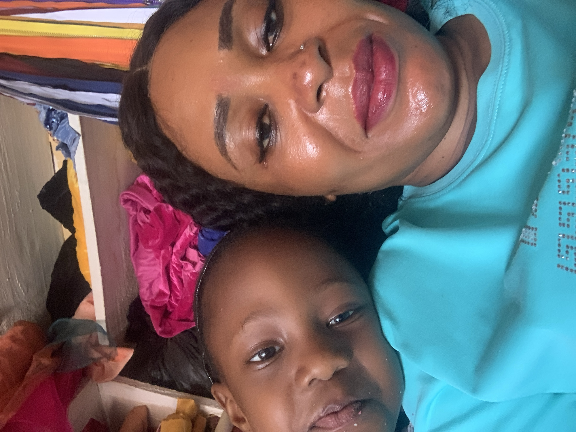 The image depicts two individuals, likely a woman and a child, in close proximity. Both individuals have dark skin and dark hair. The woman is wearing a turquoise shirt and appears to have makeup on. A nose piercing is also visible. The child appears to have remnants of food or sweets on their face. The background is cluttered with various fabrics and objects, suggesting an interior location. The location of the image is claimed to be in the city of Sabon Tasha, Nigeria. The image is oriented in a rotated manner.