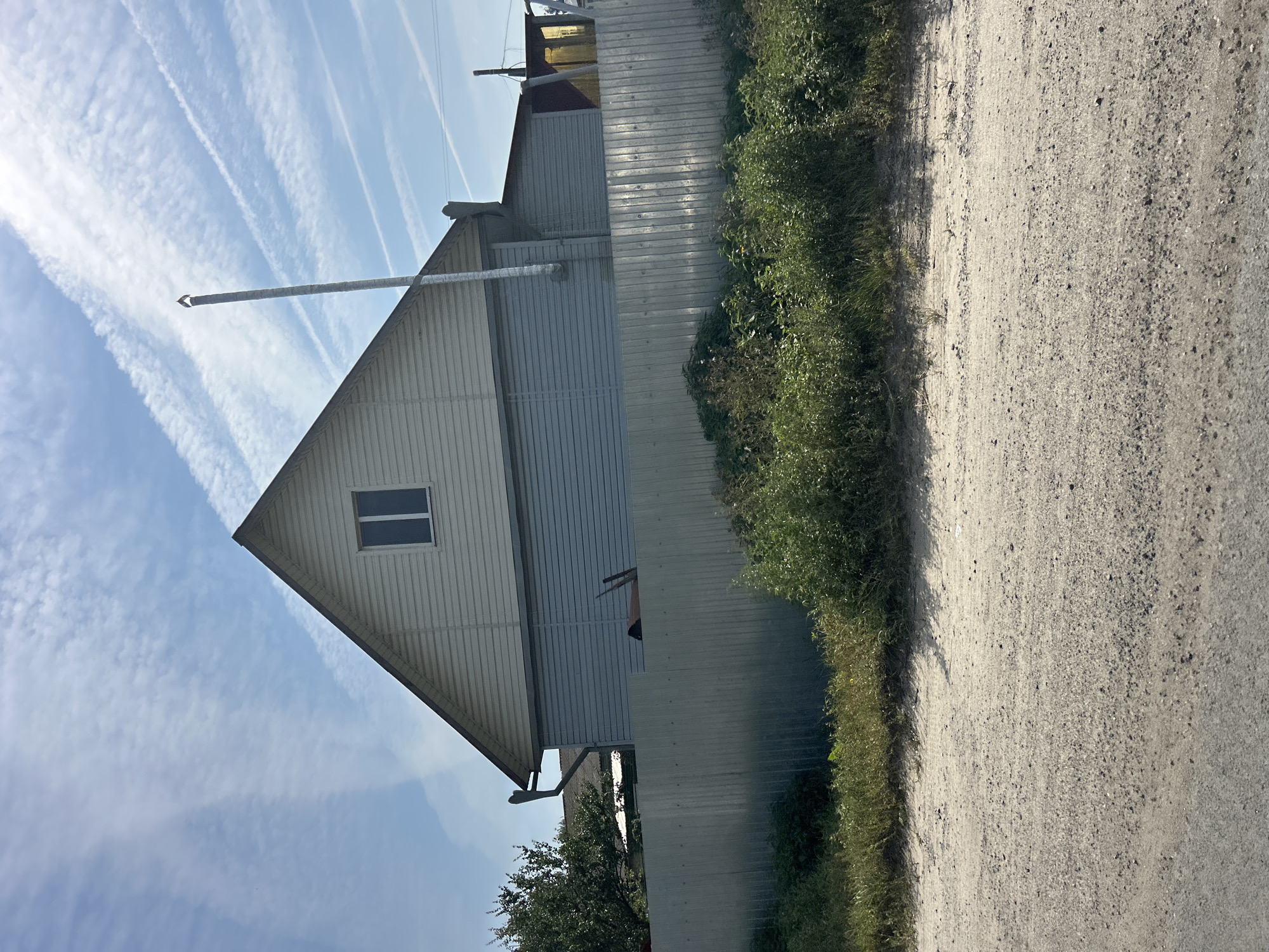 The image captures a rural scene with a house and a fence under a partly cloudy sky. The house has a light-colored, gabled roof and pale blue siding on the lower portion of the walls. A single window is visible on the upper section of the house. A tall, silver chimney extends upwards from the roof. A metal fence runs horizontally across the midground, separating the house from a strip of overgrown green vegetation in front of it. Beyond the vegetation, a dirt road occupies the foreground. The sky is blue with numerous white contrails from airplanes. The overall impression is one of a quiet, possibly remote, residential area. The scene is devoid of people or any discernible activity.