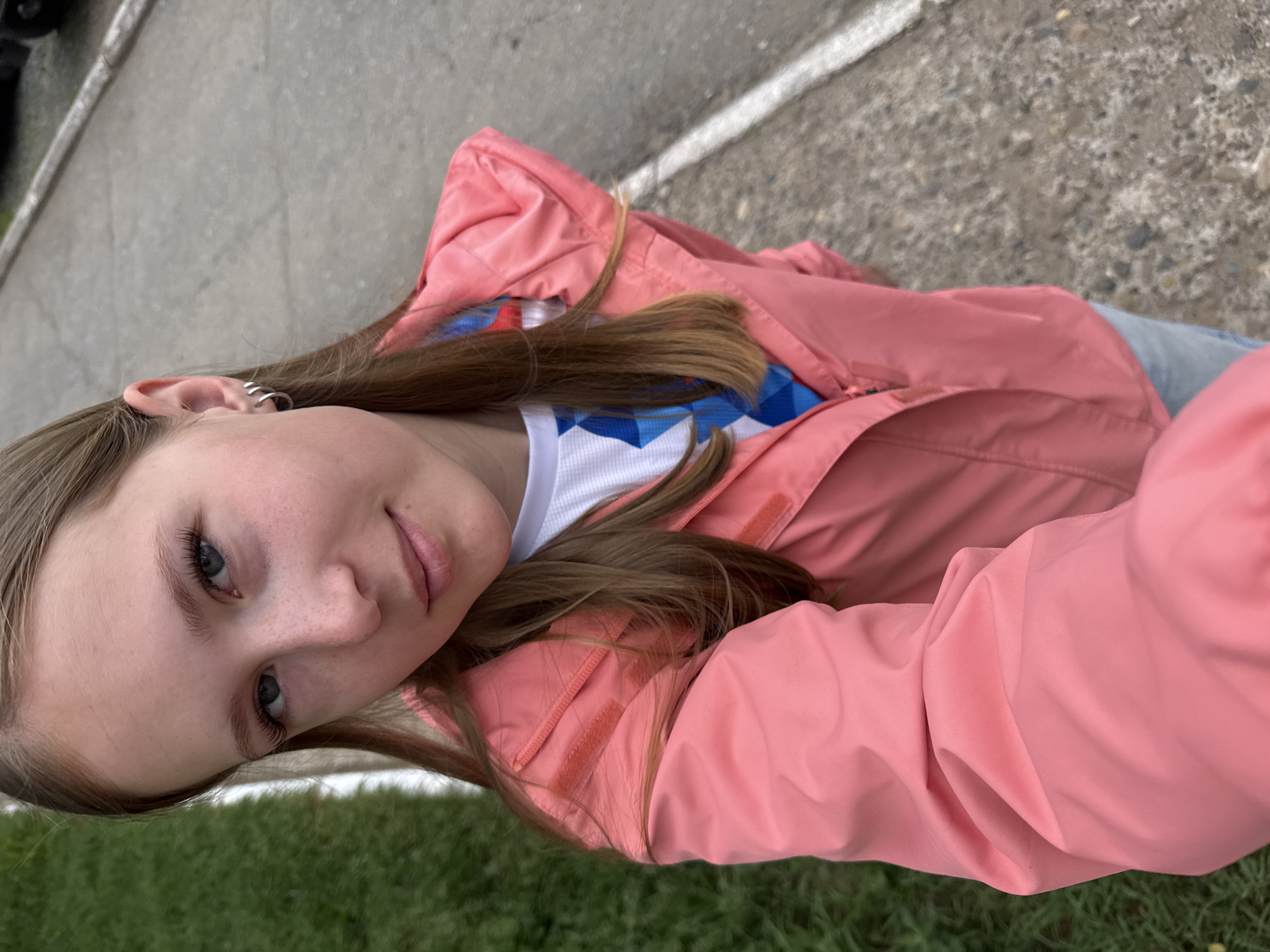 A young Caucasian girl with long brown hair, blue eyes, and freckles is taking a selfie outdoors. She is wearing a pink hooded jacket over a white t-shirt with a blue and white geometric pattern. She has small hoop earrings in her left ear. The background shows a paved surface with a white line, and a strip of green grass. The image appears to have been taken on a cloudy day. The girl is looking directly at the camera with a slight smile. The location is a street in Chaikovsky, Russia.
