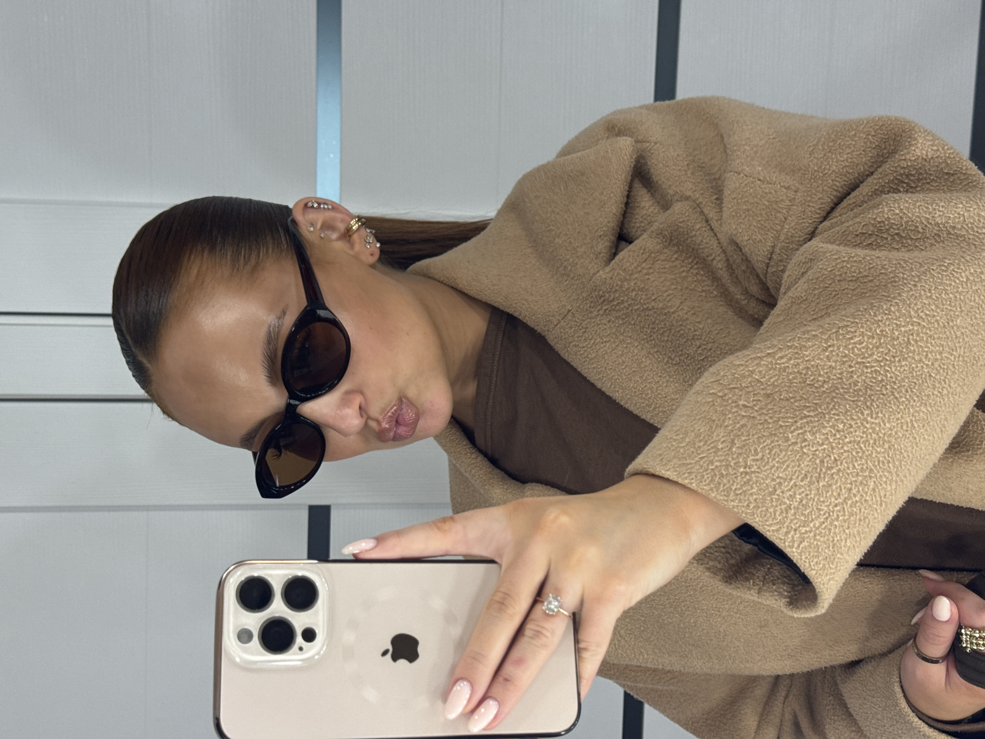 A woman in a brown coat and sunglasses is taking a selfie with a smartphone. She is wearing earrings and a ring. The background appears to be a wall with vertical panels. The location is not provided.