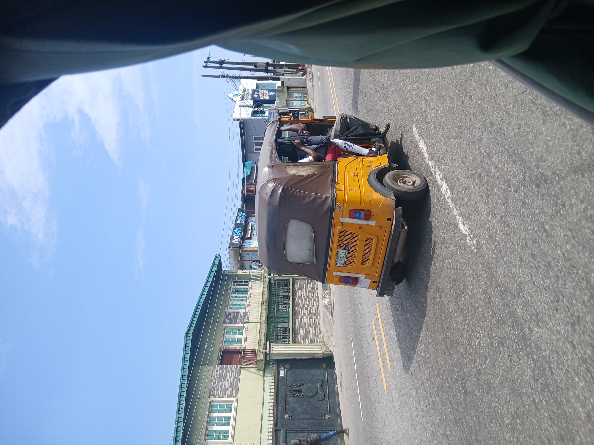 In Animashawun, Nigeria, on a bright, clear day, a yellow and brown auto-rickshaw, also known as a Keke Napep, is captured in motion. The vehicle is traveling on an asphalt road marked with yellow and white lines. The auto-rickshaw has a brown fabric roof and is carrying at least two passengers. One passenger is visible in the front, possibly the driver, wearing a white shirt and red shorts. Another passenger is seated behind, with their legs extending out of the vehicle. A third person, dressed in a dark t-shirt and blue jeans, walks on the side of the road in front of a large, dark metal gate. Behind the gate, a two-story building with a green roof and light green walls stands prominently. The building features multiple windows with white frames. To the right of the auto-rickshaw, a few shops with various signs, including one that reads "bet9ja" and another with "Vesali.com," are visible. Overhead, power lines and poles stretch across the sky, under a clear blue sky with scattered white clouds. The image appears to have been taken from inside another vehicle, as the edge of a dark green fabric is visible in the foreground.