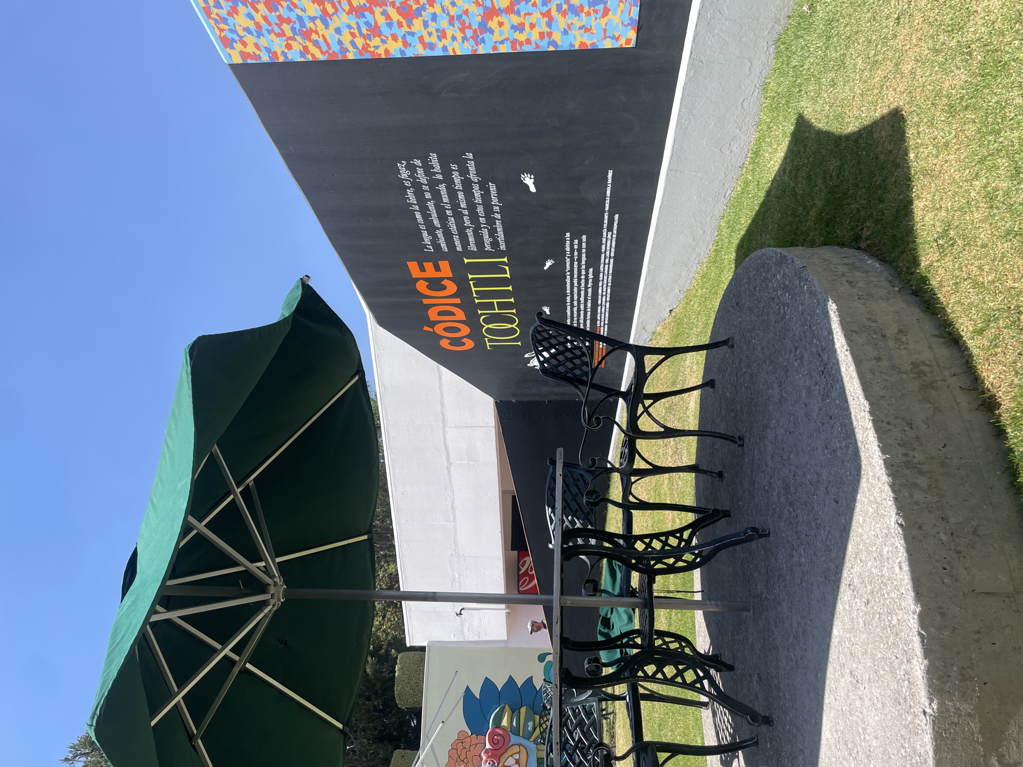 An outdoor scene in San Andrés Cholula, Mexico, depicts a seating area under a clear blue sky. In the foreground, a dark green outdoor umbrella is open, centered over a round, dark metal table with an ornate, scrolled design. Three matching dark metal chairs with similar designs are arranged around the table on a circular concrete platform. The platform is surrounded by short, well-maintained green grass.
In the mid-ground, a dark gray-black wall stretches across the frame, featuring prominent orange text that reads "CÓDICE TOCHTLI." Below this, white text in Spanish describes language, starting with "La lengua es como la liebre, es fugaz, cambiante, ambulante..." (Language is like the hare, fleeting, changing, wandering...). Small white pictograms, resembling paw prints, are also visible. The far right section of this wall is adorned with a vertical mosaic of small, irregular shapes in red, orange, blue, and yellow.
To the left of the dark wall, a white building or wall is visible, featuring a vibrant, stylized mural of a large, multi-colored creature or head with a red face, blue, and orange elements. Behind the umbrella's central pole and table, a partial view of a person wearing a light-colored cap is visible, standing in a shaded doorway or recess of the white building. A faint red "Coca-Cola" logo is discernable behind this individual. The scene is brightly lit by direct sunlight, casting distinct shadows from the umbrella and concrete platform onto the grass. No active human interactions are depicted.