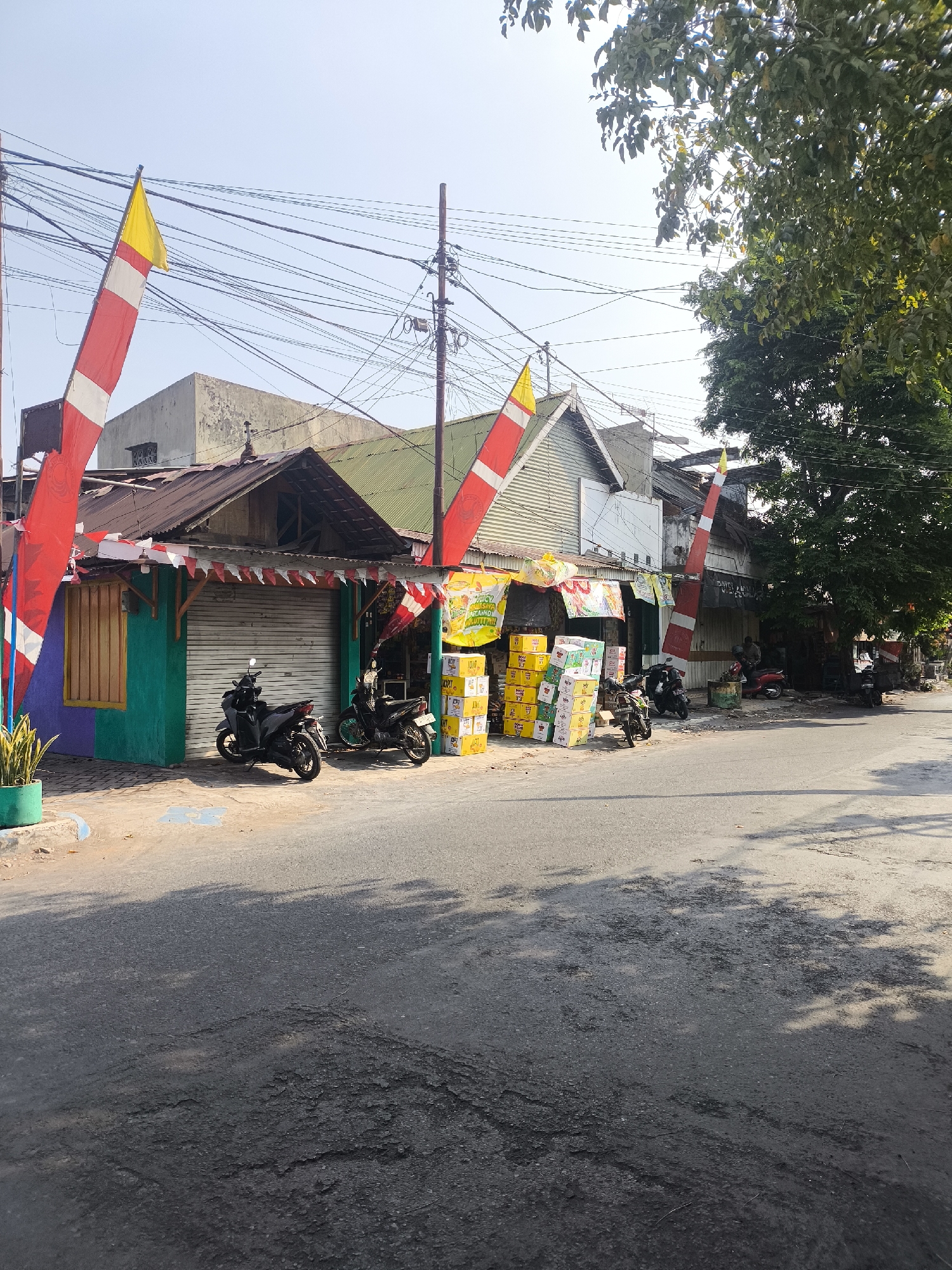 The image depicts a street scene in Lumajang, Indonesia. In the foreground, a weathered asphalt road is visible, with patches of darker, more deteriorated pavement. Sunlight casts dappled shadows of tree branches across the road. On the right side of the road, a large tree with lush green foliage overhangs the scene. Adjacent to the road are several small storefronts. One storefront on the left is painted with vibrant purple and green colors, featuring a wooden-framed window. A potted plant sits to the left of this building. Several motorcycles are parked along the storefronts. Large stacks of yellow boxes, possibly containing goods for sale, are placed in front of one of the shops. Red and white flags, characteristic of Indonesian national decorations, are prominently displayed, attached to poles and strung along the building eaves. These flags appear to be for a festive occasion. The buildings themselves have corrugated metal roofs and varying facades, suggesting a commercial area. Overhead, a dense network of electrical wires crisscrosses the sky, supported by wooden poles. In the background, the sky is clear and bright blue. A few people can be seen in the distance, near the storefronts, suggesting some activity. The overall impression is a typical street in a commercial district of an Indonesian city, likely decorated for a national celebration.