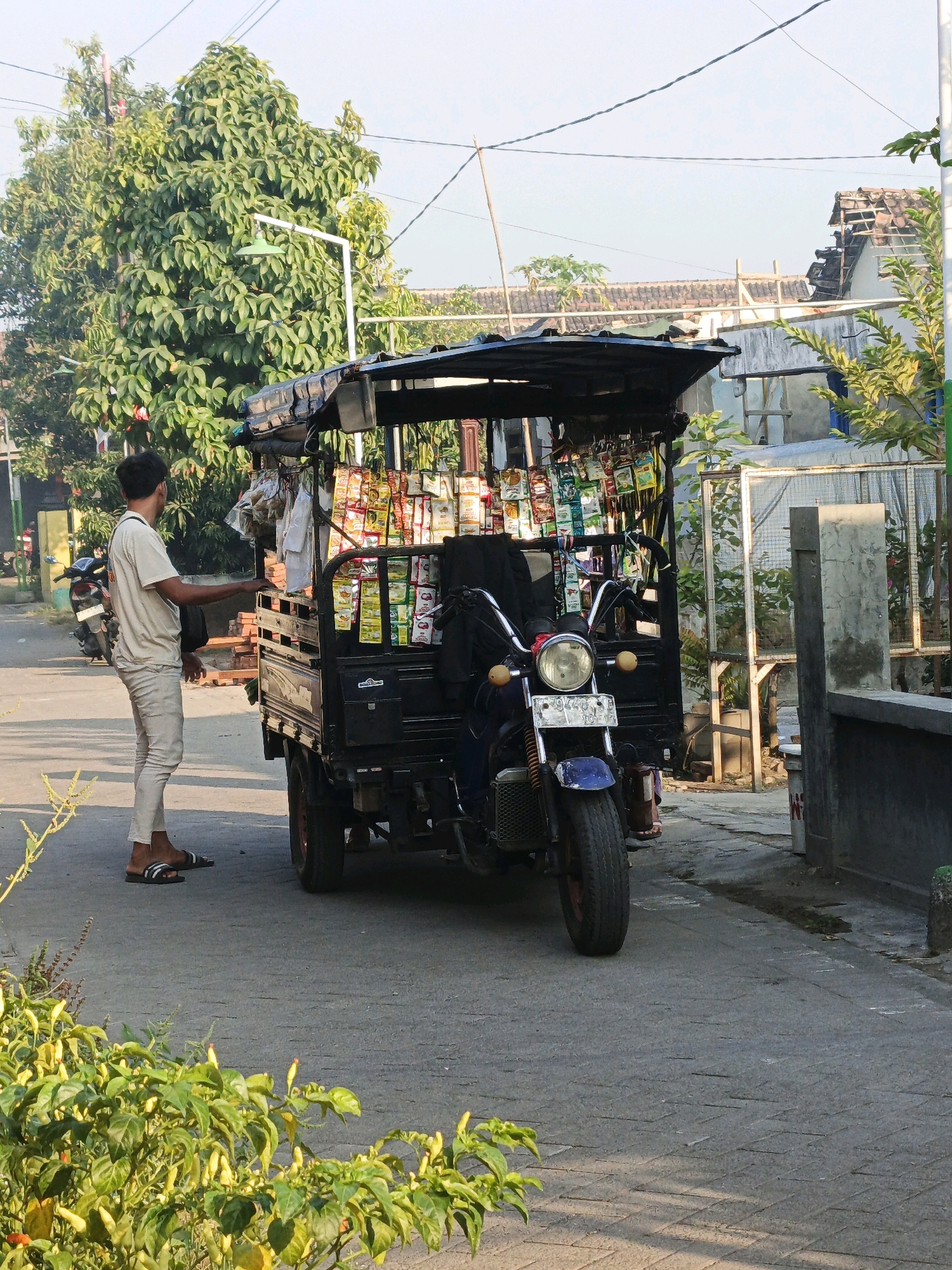 In Ketegan, Indonesia, a man in a light-colored t-shirt and pants stands beside a parked motorcycle-based food cart. The cart is loaded with various packaged goods, likely snacks or condiments, displayed on shelves under a black canopy. A white motorcycle is parked behind the man. The scene unfolds on a paved road, with houses and lush green trees lining the background under a clear sky. Sunlight casts shadows on the road and foliage, indicating it's daytime.