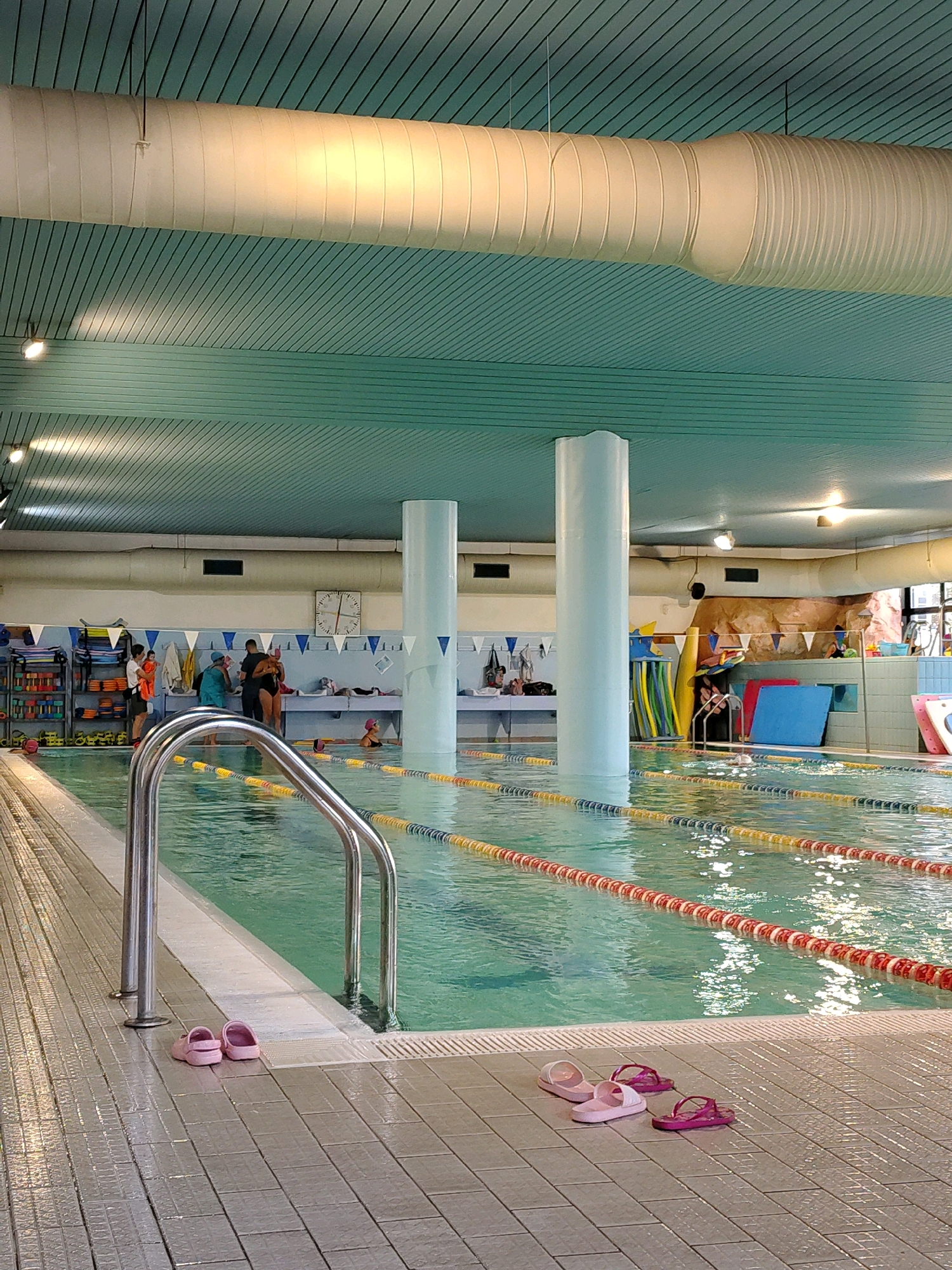 An indoor swimming pool facility is depicted. The pool is divided into lanes by ropes with buoys. The water is clear and green. The surrounding area is tiled and wet. There are several pairs of flip-flops on the tiled deck near the pool. In the background, there are people gathered near the pool's edge, some appearing to be children and adults. Equipment such as pool noodles and mats are visible on shelves and near the pool. A large, cylindrical ventilation duct runs along the ceiling, which is a light teal color with a slatted design. Two large, white cylindrical pillars rise from the water and extend to the ceiling. A clock is visible on the wall in the background. The lighting in the facility appears to be a mix of overhead fixtures and natural light.