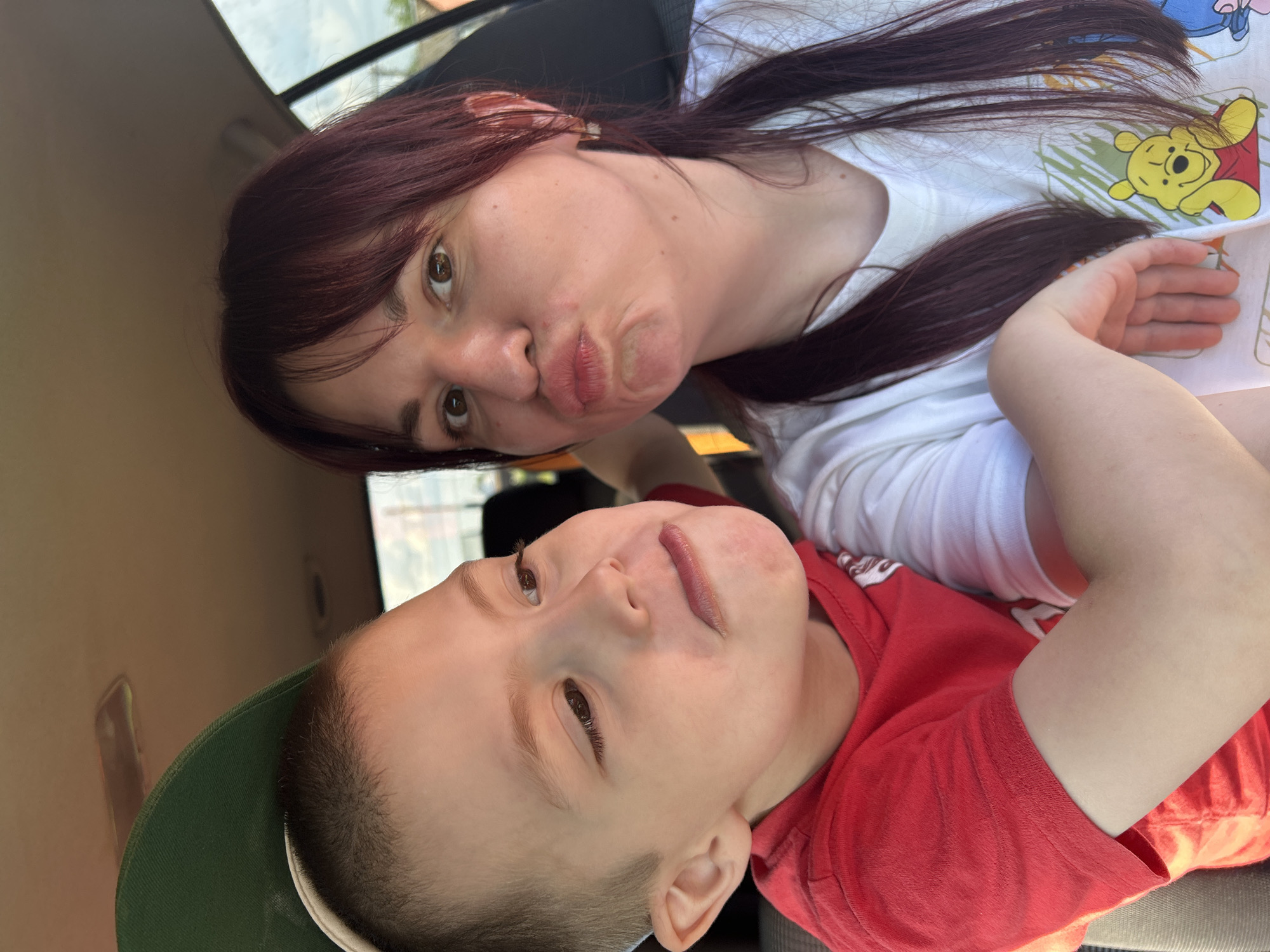 In the image, a young boy wearing a red t-shirt and a green baseball cap is positioned on the left side, looking to his left with a pout. To his right, a woman with dark hair pulled into a ponytail and wearing a white t-shirt with a Winnie the Pooh graphic is looking directly at the camera and puckering her lips. Both individuals appear to be in the interior of a vehicle, suggested by the car's ceiling and seat visible in the background. The scene is captured in a close-up, selfie-style photograph.