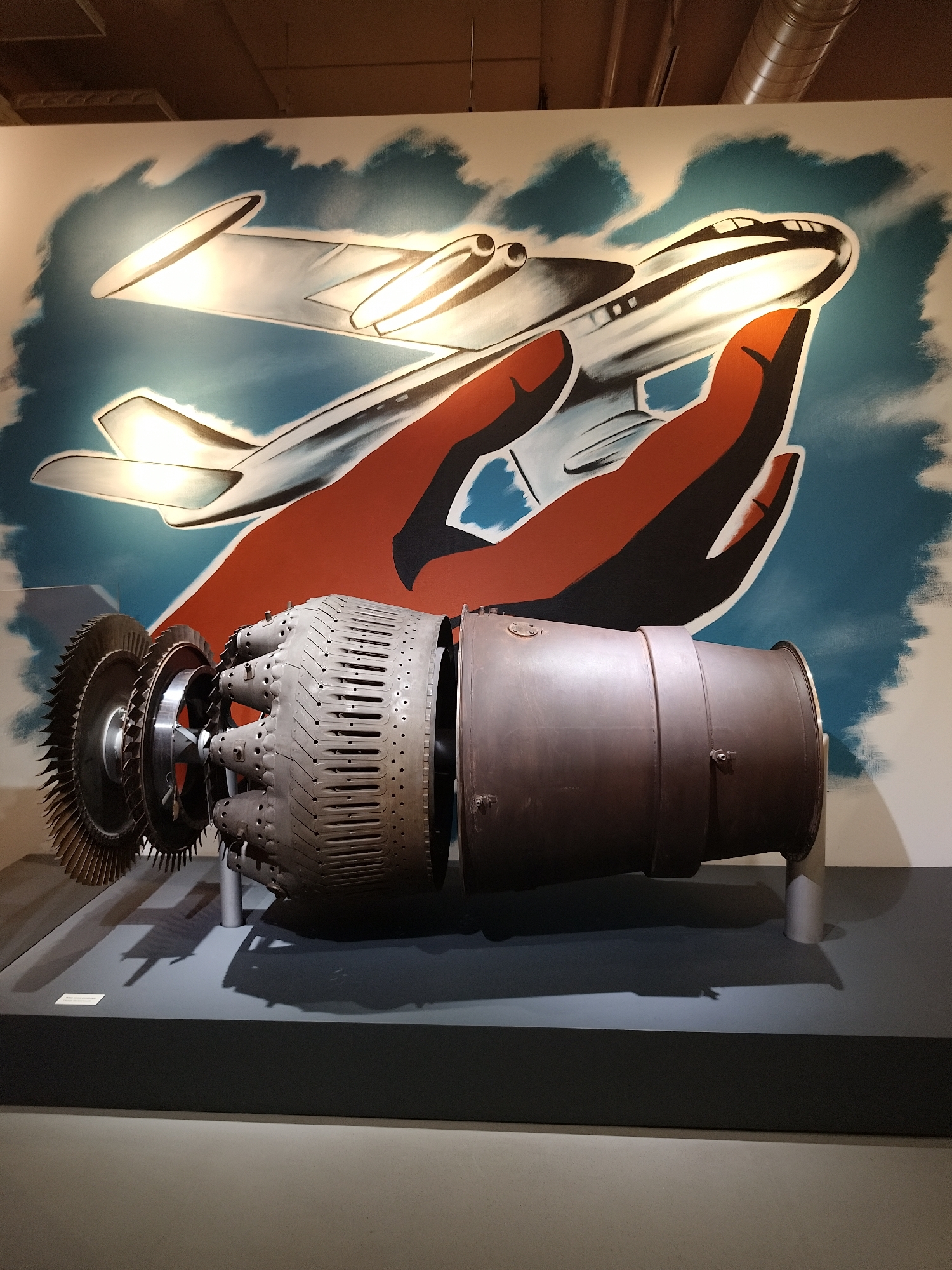 The image shows a disassembled jet engine on display in an indoor setting. Behind the engine is a mural depicting an airplane being held in cupped hands against a blue sky. The jet engine is supported by stands on a gray platform with a dark gray border. The location is Unknown, Unknown.