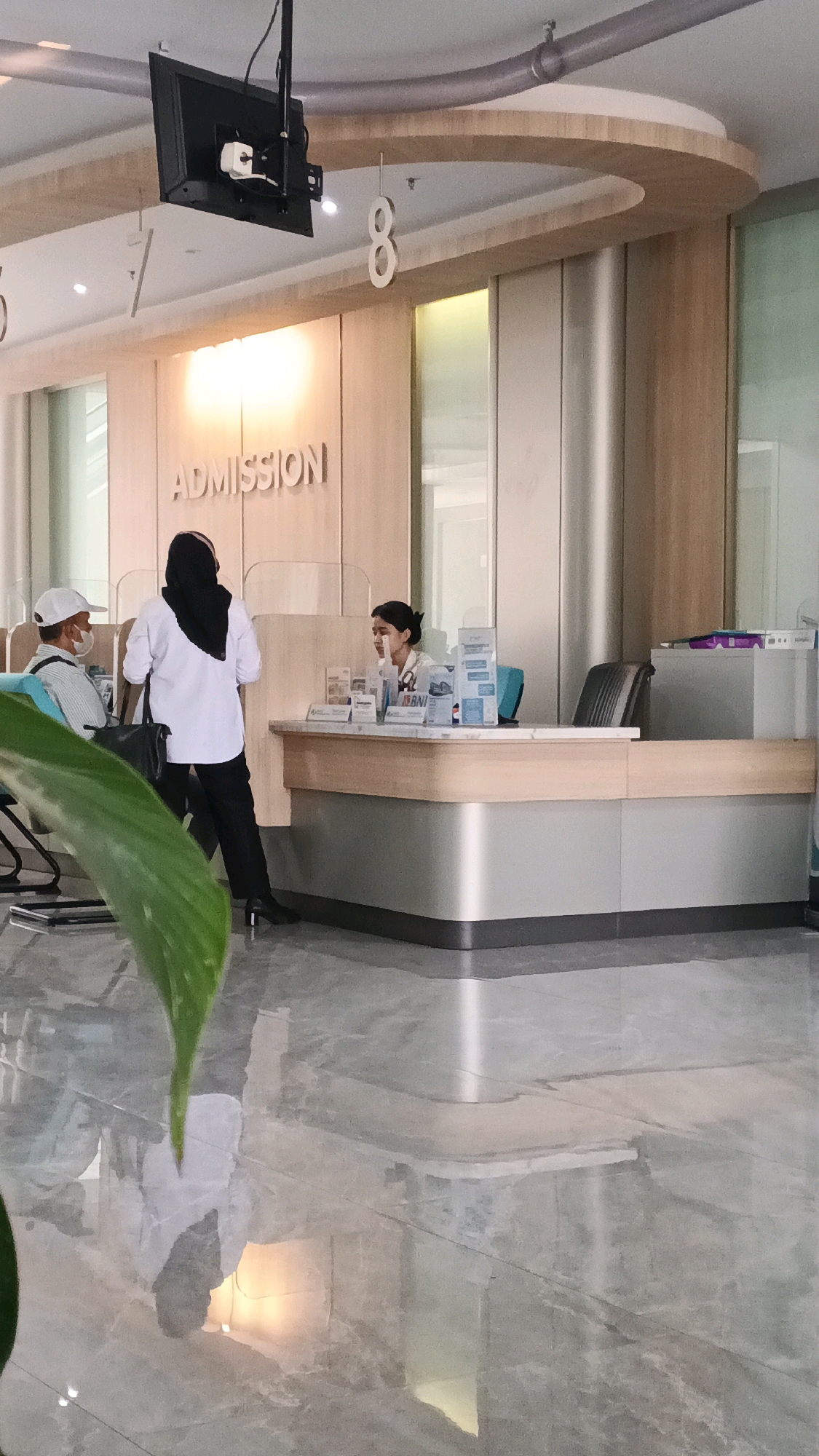A brightly lit indoor scene captures the admission area of a modern building, possibly a hospital or a bank, in Semarang, Indonesia. The walls are clad in light-colored wood panels with subtle vertical accents. Prominently displayed on the wall is the word "ADMISSION" in raised, dark letters. A sleek, curved counter, also with wooden and metallic finishes, dominates the mid-ground.
Behind the counter, a woman with dark hair pulled back is seated, wearing a light-colored uniform and appears to be interacting with a customer. To the left of the counter, a woman in a black hijab and white shirt, and a man wearing a white cap and mask, are standing. They appear to be in conversation, possibly at the admission desk.
Various brochures and informational displays are visible on the counter. Above the counter, a black monitor is suspended from the ceiling. The floor is polished and reflective, showing the reflections of the people, the counter, and the lighting fixtures. A large, green leaf in the foreground partially obscures the view, adding a touch of nature to the clean, minimalist environment. The overall impression is one of an organized and efficient service area.