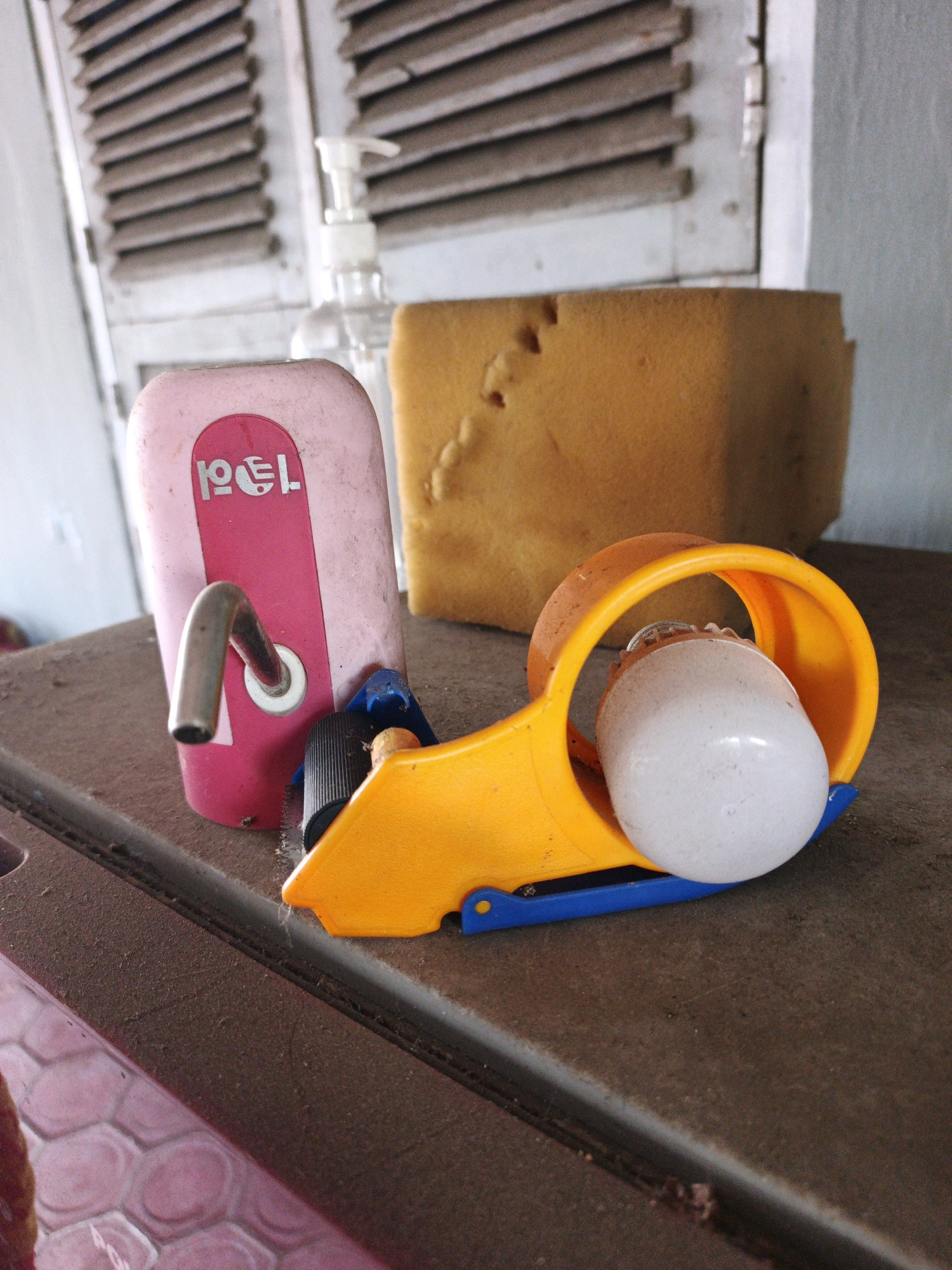The image depicts a close-up view of a dusty surface, likely a table, within a room in Ketegan, Indonesia. In the foreground, a yellow and blue tape dispenser with a white lightbulb is prominently featured. To the left of the tape dispenser is a pink water dispenser with a metal spigot and a red and white logo. Behind these items, a yellow sponge-like object rests against a white wall with louvered shutters. The overall environment appears somewhat neglected, indicated by the presence of dust on the surfaces. No people are visible in the image.