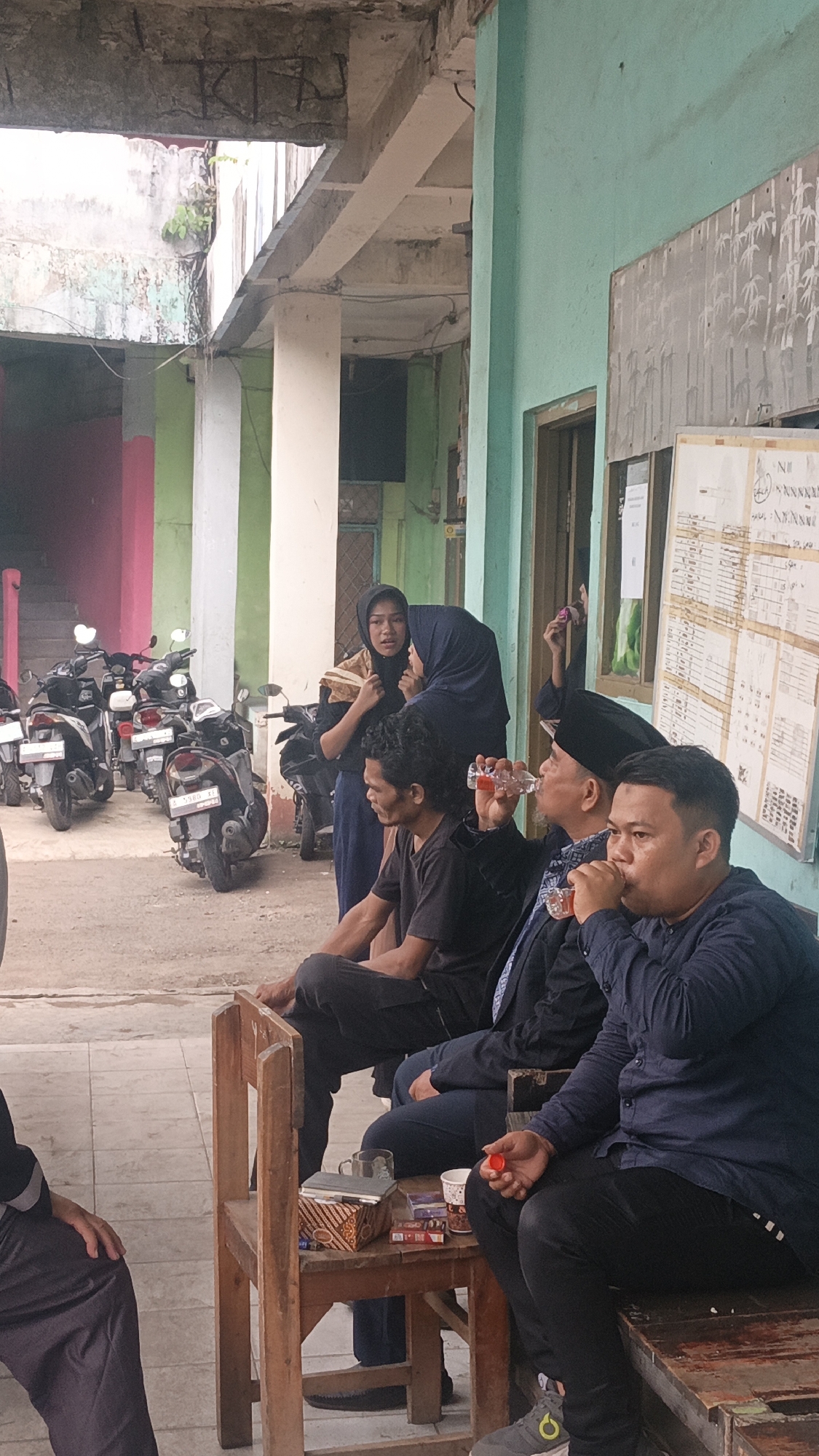 The image shows an outdoor scene at a building in Tigaraksa, Indonesia. In the foreground, three men are seated on wooden benches. The man on the far right is wearing a dark blue long-sleeved shirt and black pants, and is holding a red bottle cap. The man in the middle is wearing a dark suit jacket over a white and blue patterned shirt and black pants, and is drinking from a clear bottle with a red cap. The man on the far left is wearing a dark t-shirt and black pants, and appears to be looking forward. Between the two men on the left is a small wooden table with a box of tissues, several packs of cigarettes, a mug, and a small plastic cup with a red lid. In the background, two women are standing, one wearing a dark headscarf and a tan and dark garment, and the other wearing a dark blue headscarf. Several motorbikes are parked behind the individuals. The building has a light green exterior wall and a covered walkway with concrete pillars. A bulletin board with papers is attached to the wall. The ground is paved with square tiles.