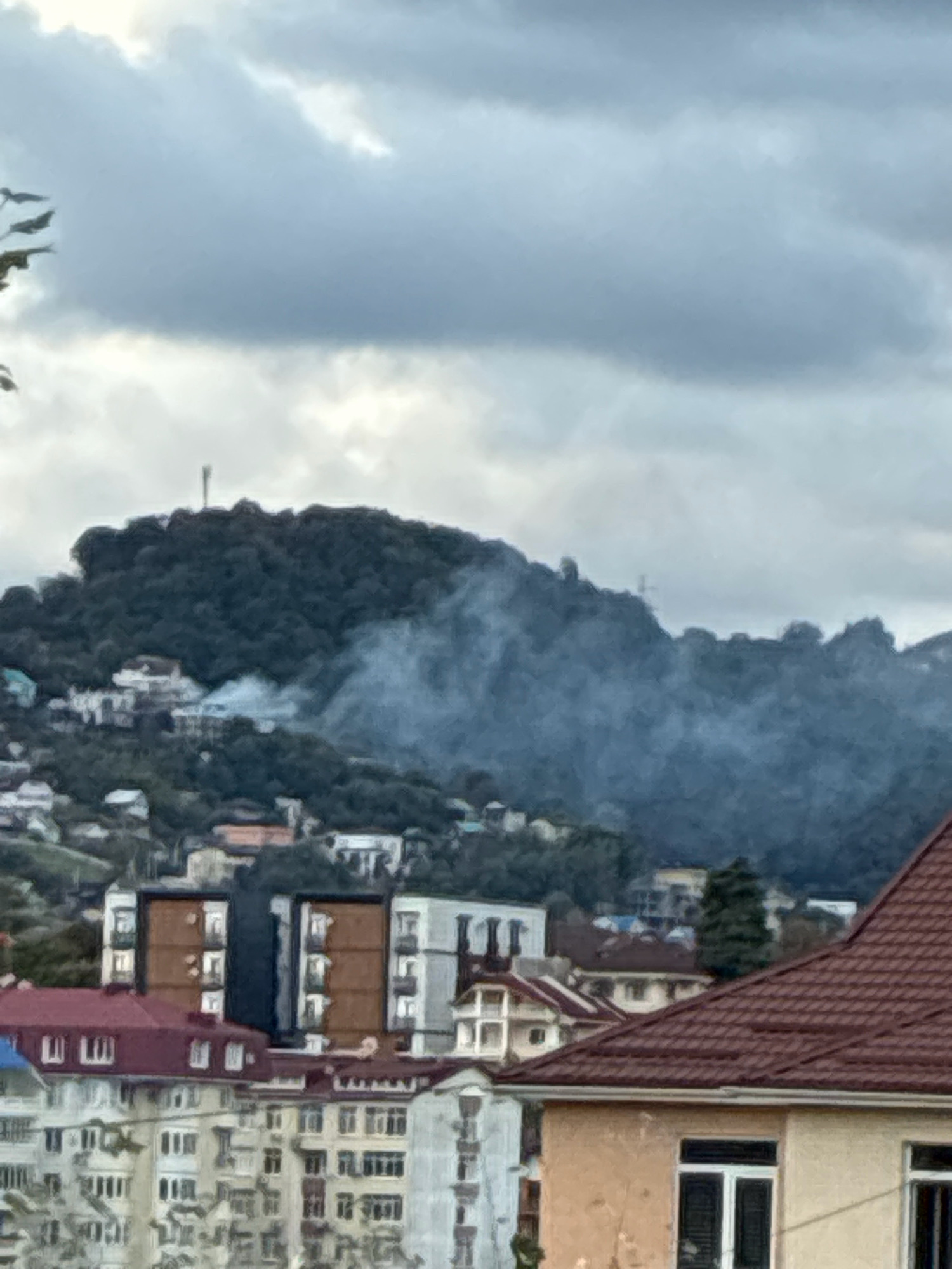 The image depicts a cloudy sky over a cityscape in Sochi, Russia. In the middle ground, a densely forested hill is visible, with a communication tower at its peak. A plume of white smoke rises from the trees on the side of the hill, indicating a fire or controlled burning. Scattered houses and buildings are nestled on the lower slopes of the hill and extend into the foreground. In the foreground, multi-story apartment buildings with red and light-colored facades dominate the view, some with visible windows and balconies. The overall atmosphere is overcast and somewhat hazy.