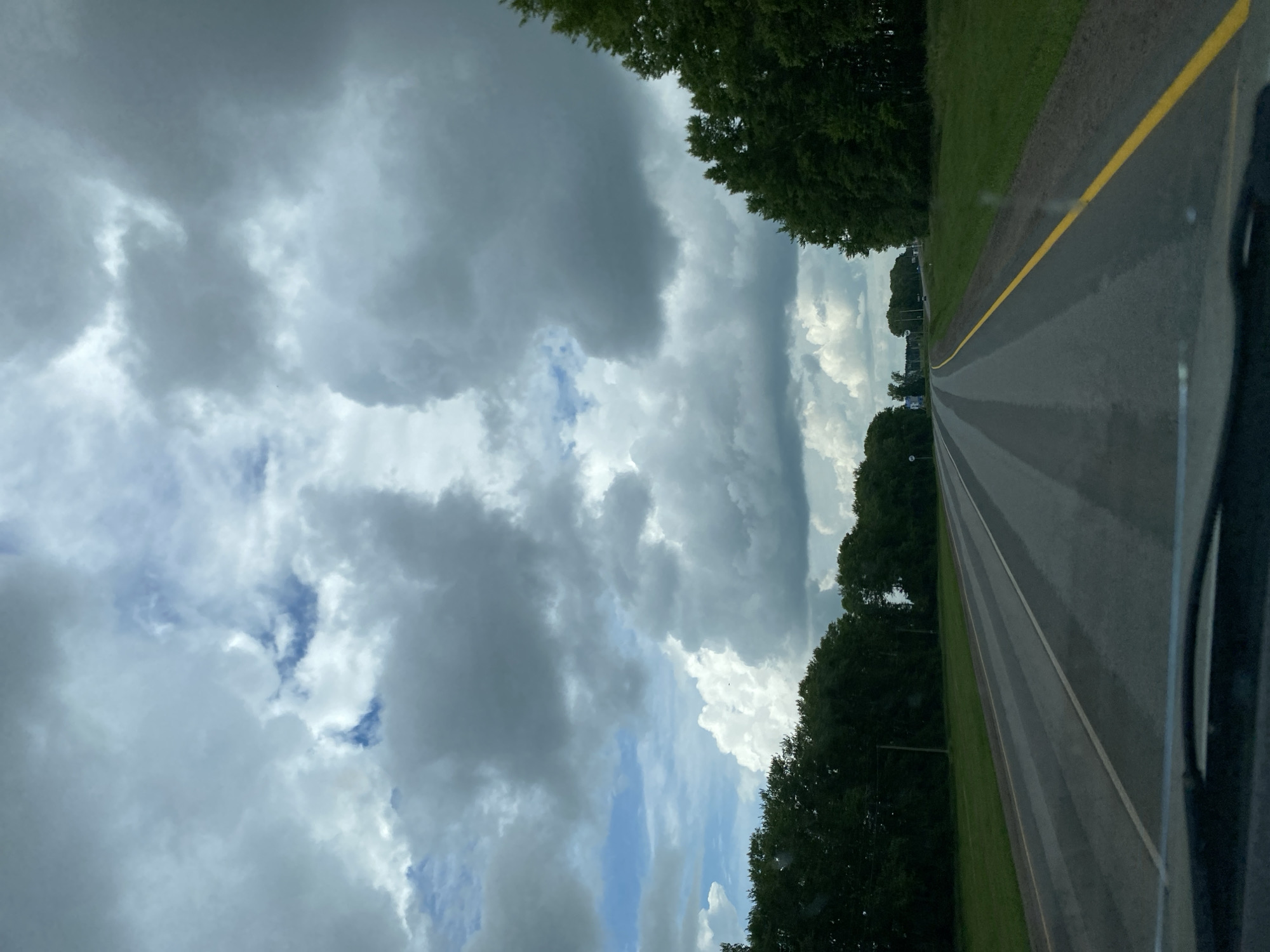 A wide-angle, eye-level, first-person perspective captures a view from a vehicle driving on a two-lane asphalt road. The road is lined with a bright yellow stripe on the right and a white stripe on the left, with lighter, striped markings on the road surface itself, possibly from recent rain. The road cuts through a rural landscape, with dense, dark green trees lining both sides of the road. The sky is filled with dramatic, grey and white cumulonimbus clouds, with patches of blue sky visible. The overall impression is that of driving through a slightly overcast, possibly humid, rural environment. No people or specific landmarks of Kushnarenkovo, Russia are identifiable.