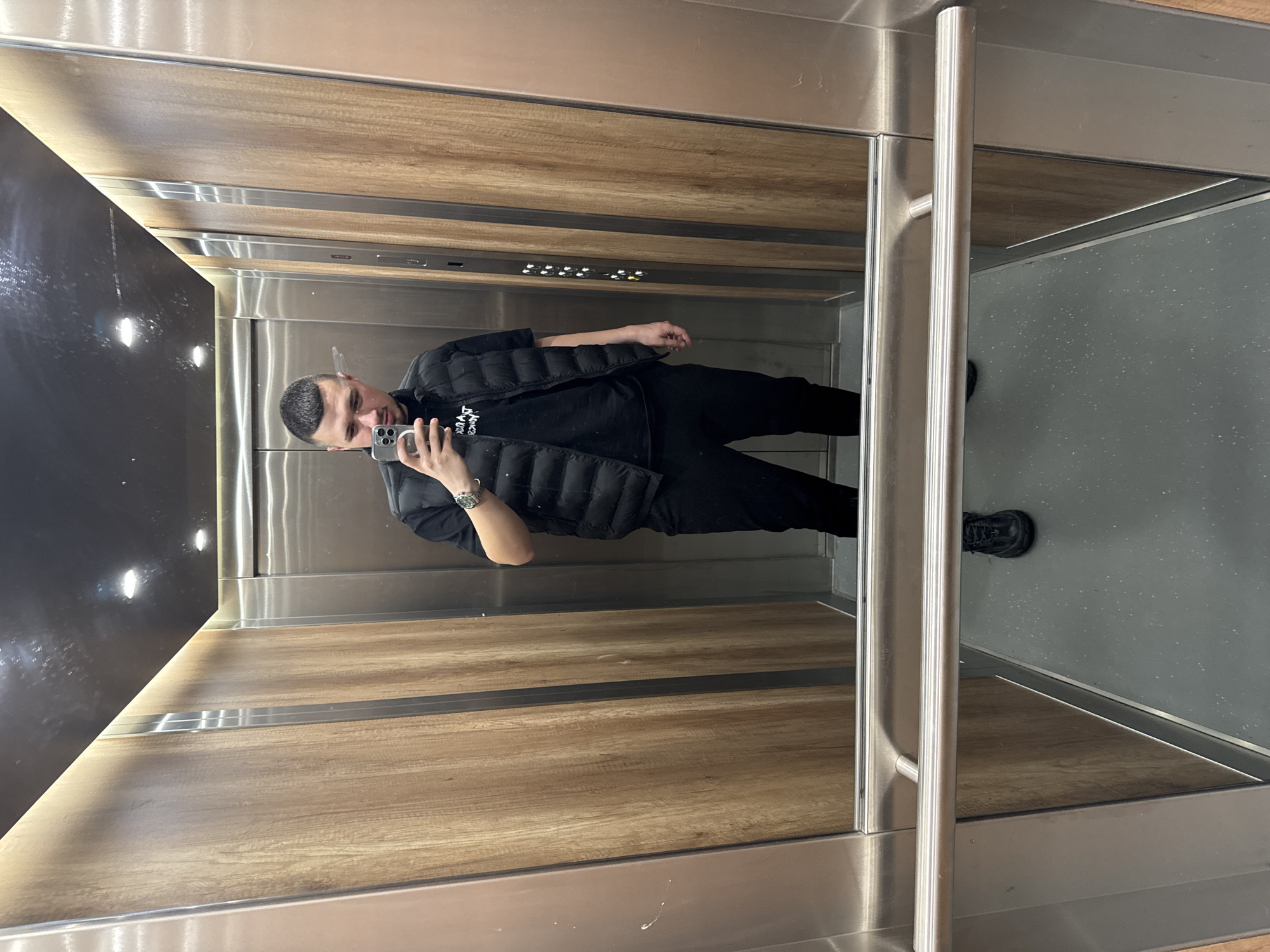 The image shows a man taking a selfie in an elevator. The elevator's interior features wood paneling on the side walls and a gray floor. A control panel with buttons is visible near the top. The man is wearing black clothing and a vest. A watch is visible on his left wrist. The location is Sofia, Bulgaria, but there is no information in the image to confirm the location.