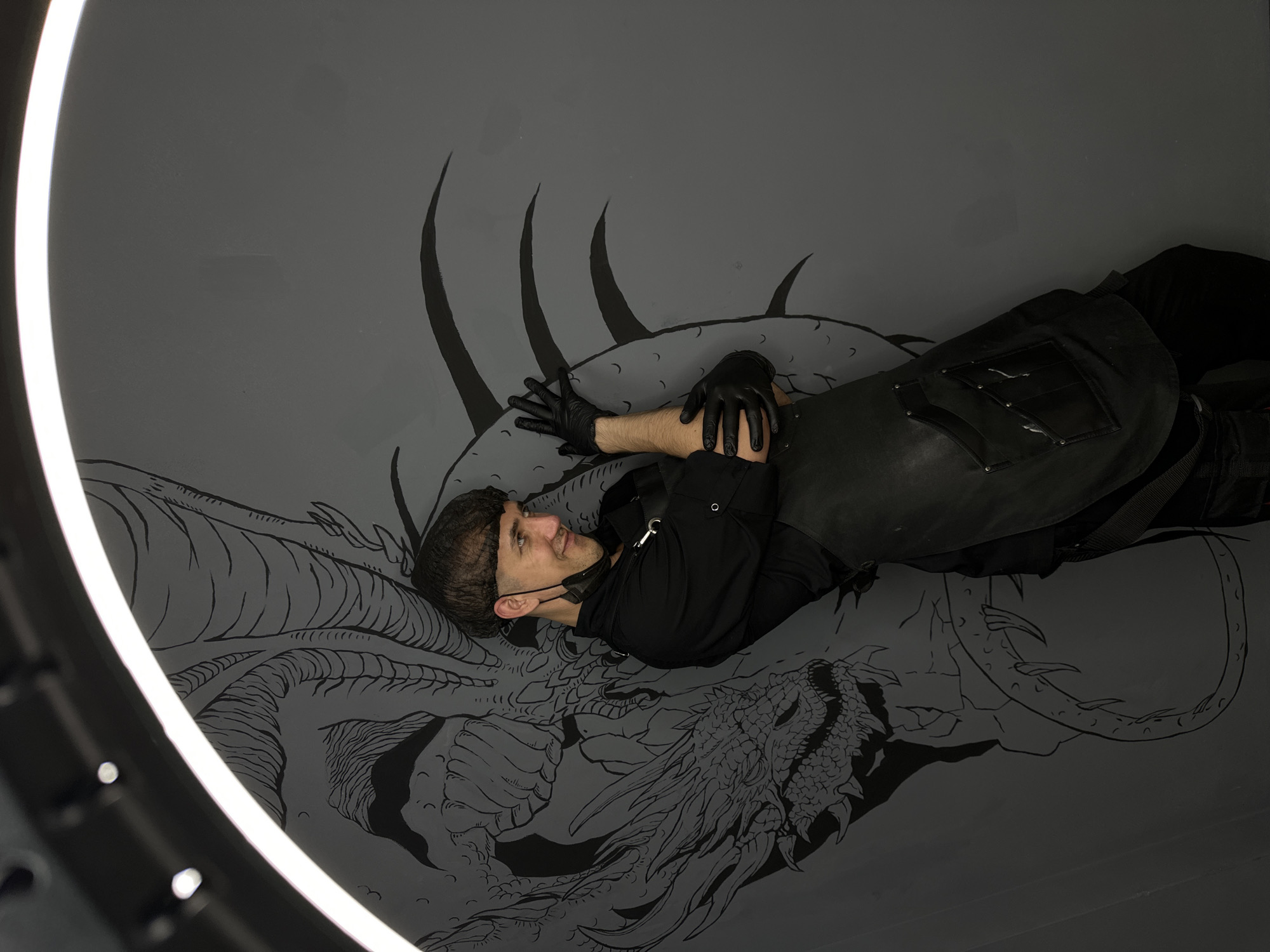 A man wearing a black shirt, black apron, black gloves, and a hair net smiles and poses provocatively against a grey wall adorned with a large, black line-drawing of a dragon. The man's left arm is bent at the elbow, with his hands resting on his chest. His right arm is also bent, with his hands near his face, as if he is admiring himself. The dragon mural appears to be the work of a local artist in Saratov, Russia.