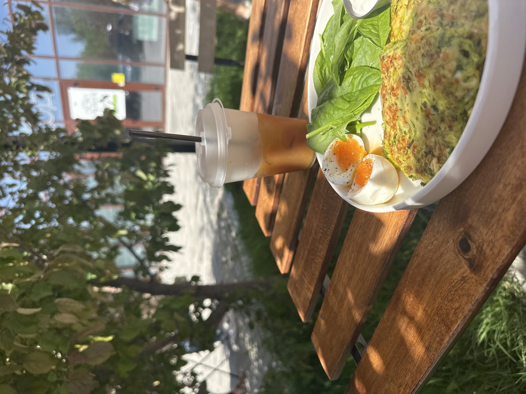 The image captures a meal and drink set on a wooden slatted surface, likely a table or bench, set outdoors, possibly in the city of Novosibirsk, Russia.
On the wooden surface, a clear plastic cup with a lid and black straw contains a layered drink, the bottom layer being light-colored and the top layer being brown, possibly a milk-based coffee drink. Next to the drink, there is a white plate containing a portion of green vegetable fritters or patties. Adjacent to the fritters are some fresh spinach leaves. Two halves of a boiled egg are also present on the plate, seasoned with black pepper. A dollop of white sauce is present, likely sour cream.
The surrounding environment appears to be outdoors. Lush green foliage, possibly from trees or shrubs, is visible around the table. A glimpse of a building is seen in the background, but it's partially obscured by the foliage. The building seems to have glass windows and doors. The foreground and background suggest an urban setting with greenery. The scene is well-lit, indicating it is likely daytime.