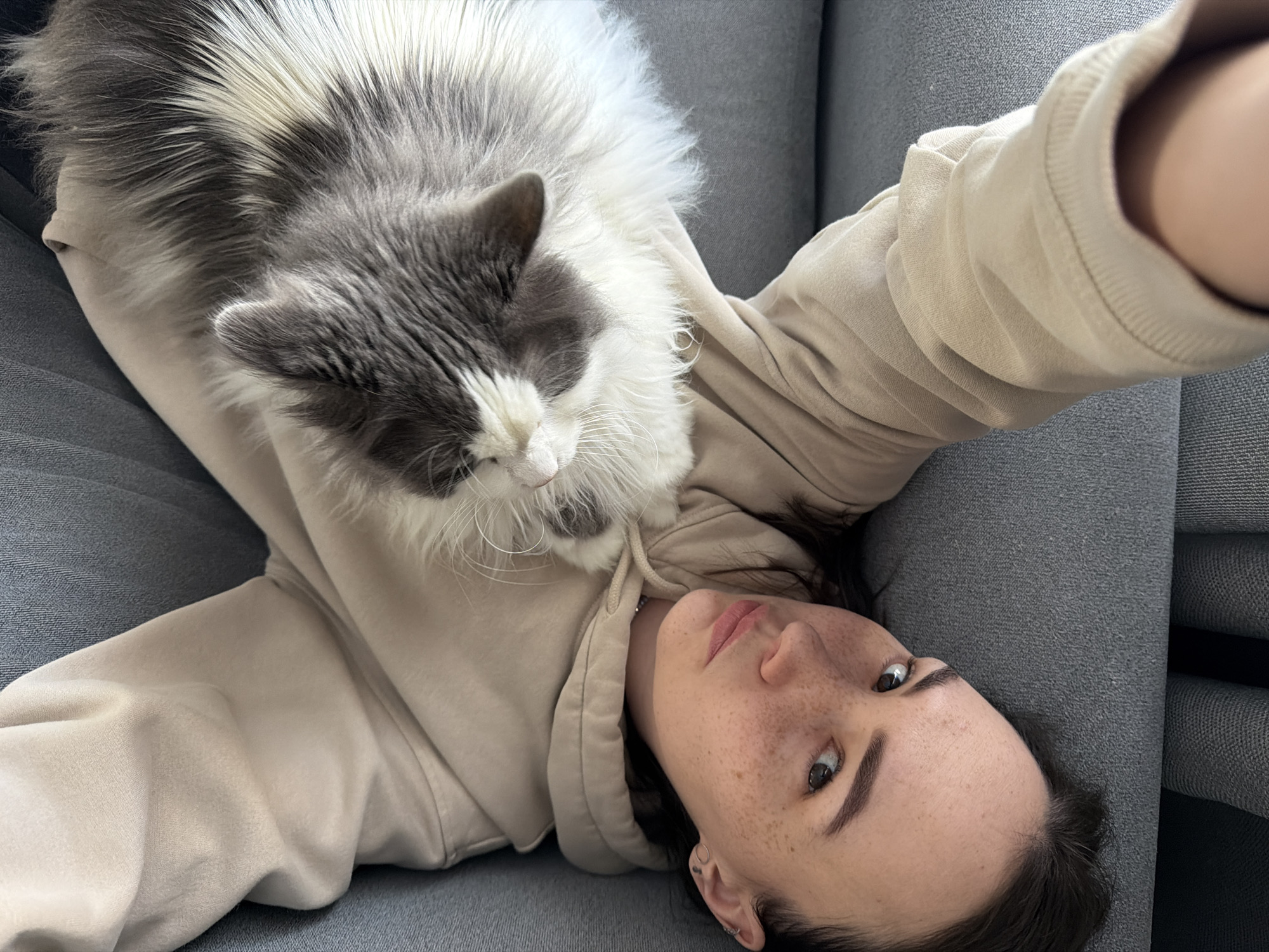 An overhead shot captures a young adult woman lying on her back on a grey fabric sofa, with a fluffy cat resting on her chest, in what appears to be a home in Chaikovsky, Russia. The woman wears a light beige hooded sweatshirt. Her dark hair is spread out, and her face, characterized by visible freckles and dark eyebrows, looks directly towards the camera with a neutral, relaxed expression. She has a subtle pinkish tint on her lips.
Perched comfortably on her upper body is a long-haired cat with prominent grey and white fur. Its head is turned slightly towards the woman's shoulder, and it appears calm and serene, possibly dozing. The setting is indoors, with the textured grey upholstery of the sofa forming the background. The scene depicts a moment of quiet companionship and relaxation. There is no visible text in the image.