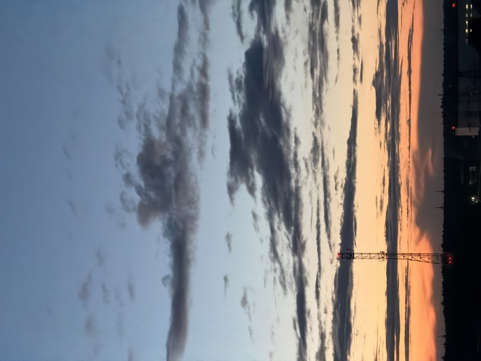An outdoor, full shot captures a cell tower against a twilight sky. The sky is a gradient of colors, transitioning from a pale blue at the top to a warm orange and yellow hue near the horizon, indicating the presence of sunset or sunrise. Scattered clouds, varying in shade from dark gray to light white, are visible across the sky. The cell tower, a tall, slender metal structure, stands prominently in the center of the frame, illuminated by red lights at its apex and base. Below the tower, a silhouetted landscape of buildings and trees suggests an urban or semi-urban environment. The overall scene depicts a late evening or early morning view in an unspecified city location.