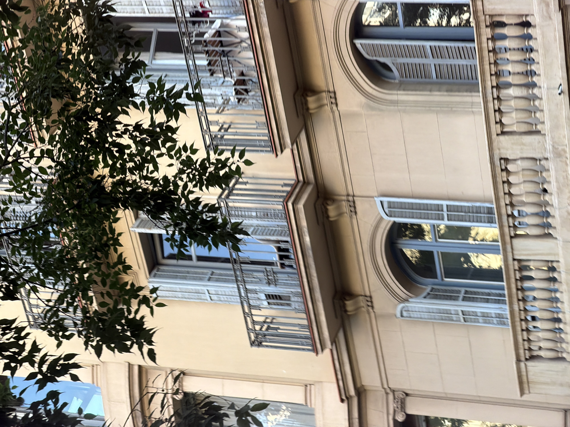 The image shows a close-up of a beige building facade in Barcelona, Spain, with balconies and windows visible. Green leaves from an overhead tree partially obscure the view. The balconies have decorative railings and some outdoor furniture is visible on one of them. The windows have light blue shutters, some of which are open. The architectural style suggests an older, possibly residential, building. The lighting indicates it might be daytime, with sunlight reflecting off the windows. No people are clearly visible, and there are no specific actions or interactions depicted.