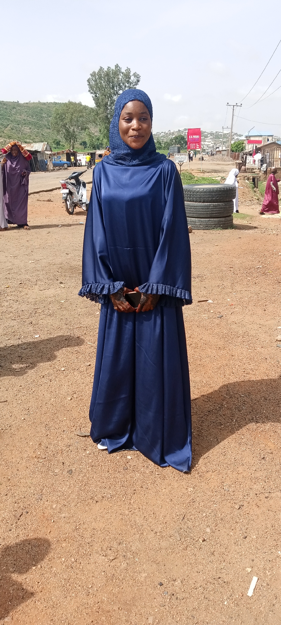 The image depicts a woman standing in what appears to be a street in Naraguta Hausawa, Nigeria. She is wearing a long, dark blue dress with lace trim at the wrists and a matching headscarf. Her hands are clasped in front of her, holding a phone.
In the background, there is a dirt road, various buildings, and a motorcycle. To the left, a person in a purple robe stands near the road. Behind the woman, there are stacked tires, and a sign with text is visible. Another person in a maroon robe is walking to the right. The landscape suggests a somewhat urbanized area with hills in the distance.
