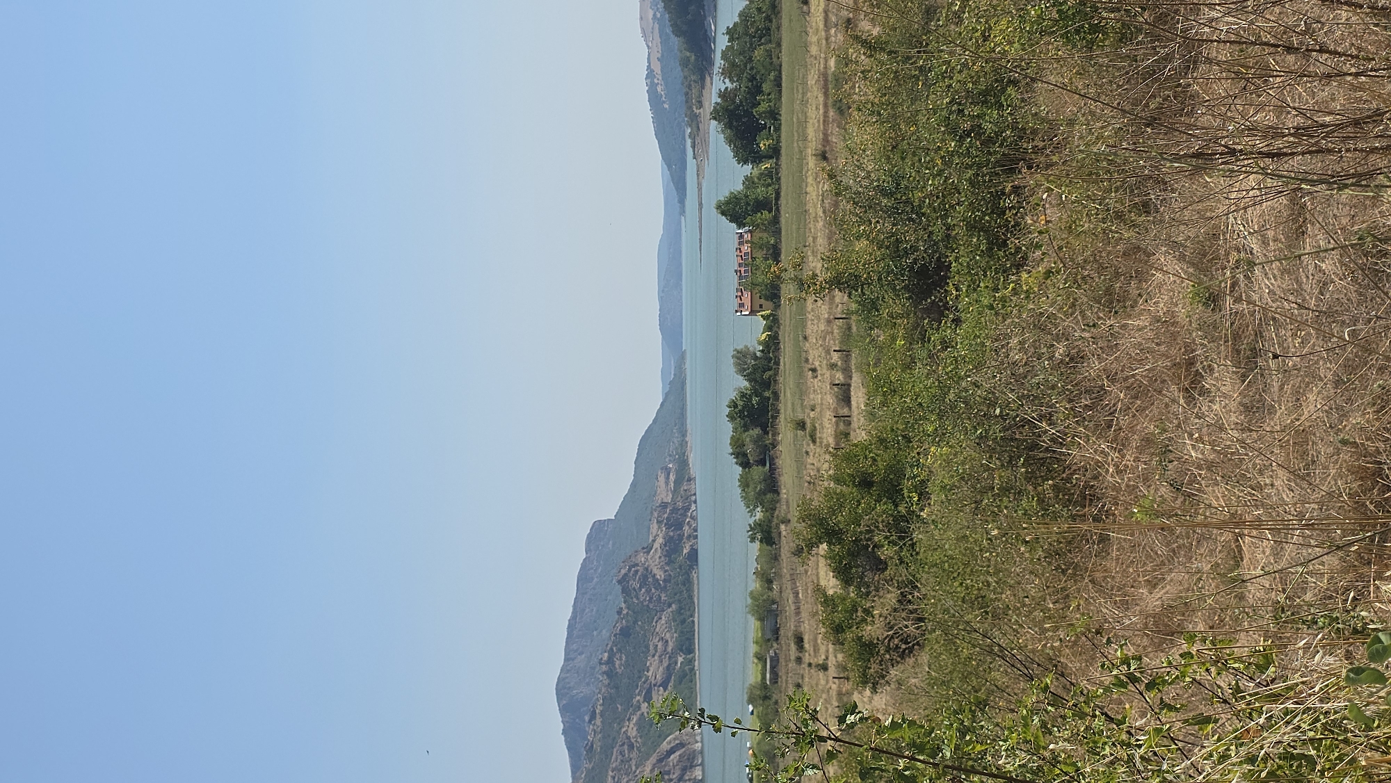 The image captures a serene landscape under a clear blue sky. The foreground is dominated by dry, tall grass and scattered green foliage, suggesting a warm, arid environment. In the mid-ground, a tranquil body of water, possibly a lake or reservoir, stretches across the frame, reflecting the clear sky. Lush green trees line the banks of the water, creating a stark contrast with the dry foreground. Further in the distance, rolling hills and rugged mountains form the backdrop, their earthy tones blending with the natural hues of the landscape. A cluster of buildings with red roofs is visible on the right bank of the water, nestled amongst the trees, hinting at a nearby settlement. The overall scene depicts a peaceful, natural setting with human presence indicated by the buildings in the distance. The location is identified as Shiroko Pole, Bulgaria.