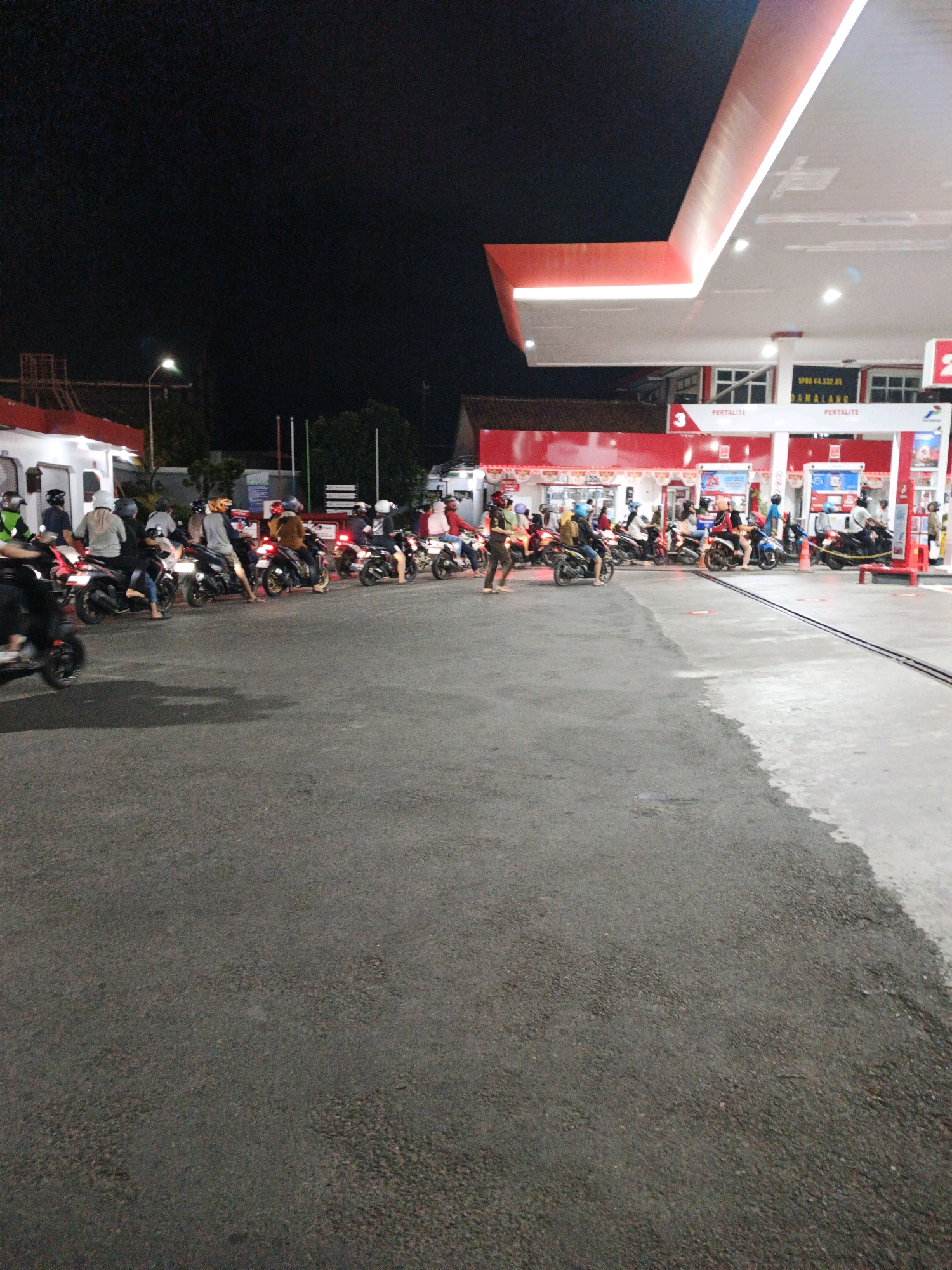 This image depicts an outdoor scene at a Pertamina gas station in Cilacap, Indonesia, at night. Several motorcycles are lined up, with riders sitting on them, presumably waiting to refuel. The gas station canopy, illuminated by bright white and red lights, extends over the area. The ground is paved with asphalt in the foreground and concrete in the background, where the gas pumps are located. In the middle ground, there are multiple rows of motorcycles and riders. The overall atmosphere is that of a busy, late-night service station.