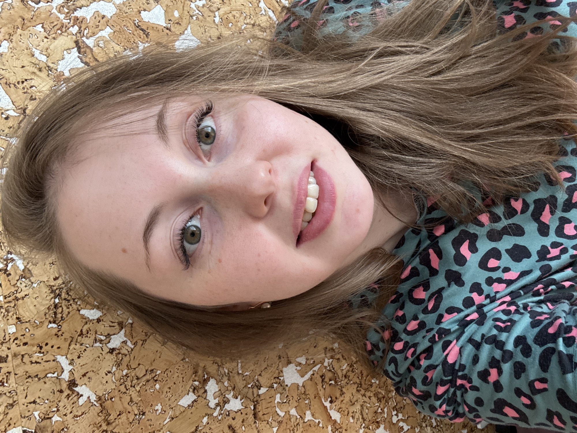 A young woman with light brown hair and fair skin is depicted in a close-up shot, angled as if she is lying down and looking upwards at the camera. Her head is tilted, with her hair fanning out around her. She has bright, light green eyes with prominent dark eyelashes and well-defined eyebrows. A subtle, pleasant smile plays on her lips, revealing her upper teeth. Her lips are colored with a soft pinkish-mauve tone, and faint freckles are visible across her nose and cheeks. She is wearing a garment with a distinctive teal or aqua background patterned with black and pink leopard spots. The background behind her is a textured surface that resembles corkboard, featuring a light brown, tan, and white fragmented design. The lighting is bright and even, suggesting an indoor setting during the day or under artificial light. There is no visible text in the image.