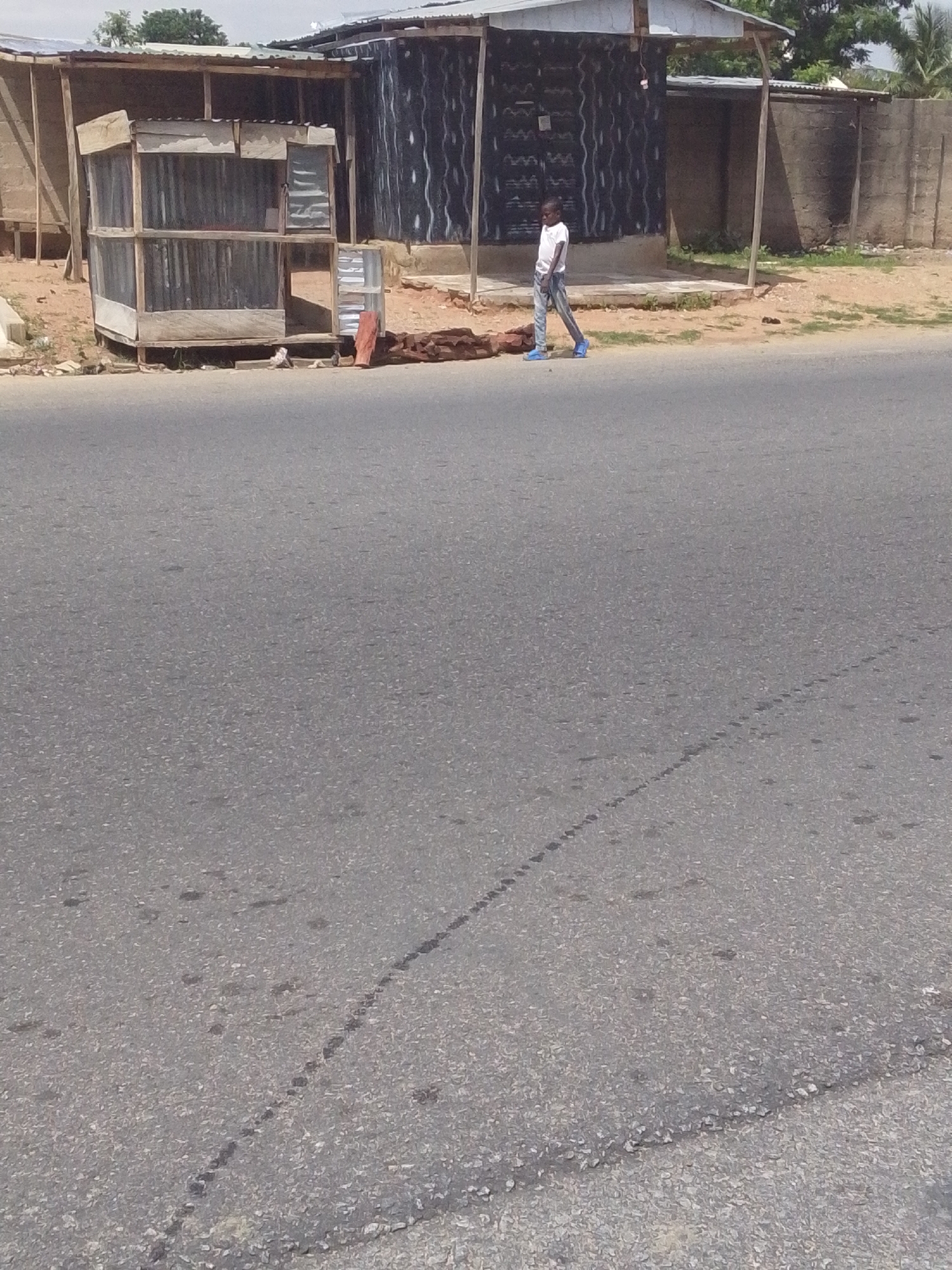 A young man in a white t-shirt and blue jeans walks on a dirt path next to a roadside kiosk made of corrugated metal and wood. He is wearing blue shoes. Behind him is a building with a black and white wavy pattern on its side, and a grey concrete wall. The road in front of the kiosk is paved with asphalt and shows tire marks. The scene is located in Jalingo, Nigeria.