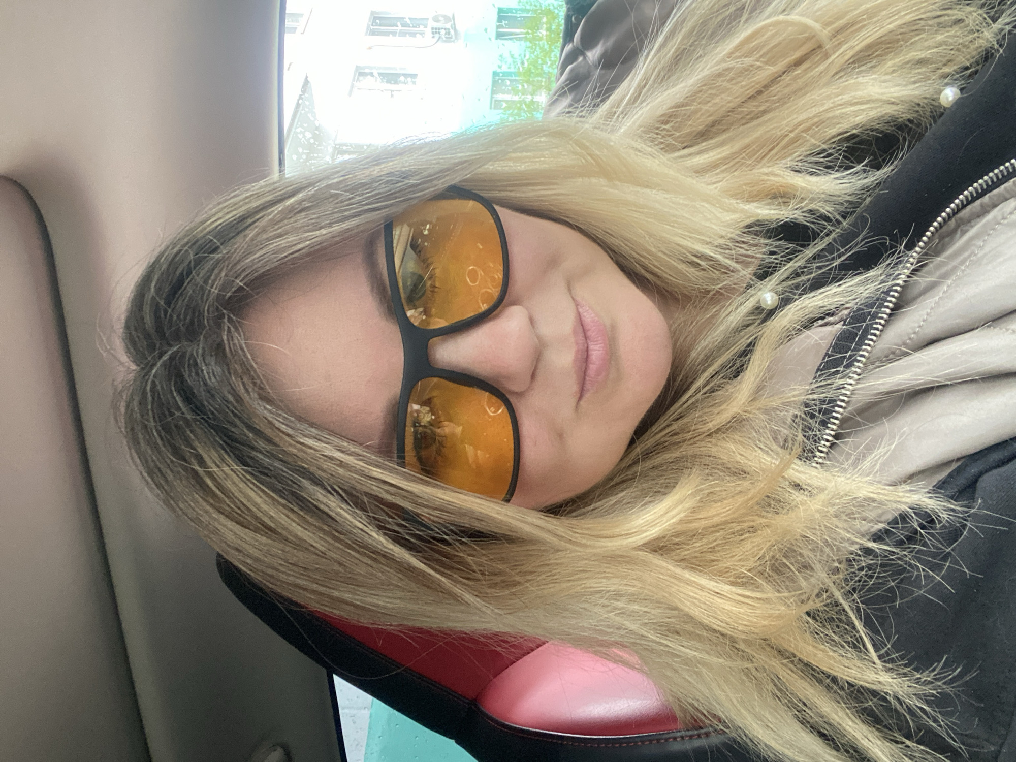 A woman with long blonde hair and dark-framed yellow-tinted sunglasses is in a vehicle. She wears a zip-up jacket. Outside the window, buildings are visible, but the location is not discernible as Chaikovsky, Russia, from the image alone.