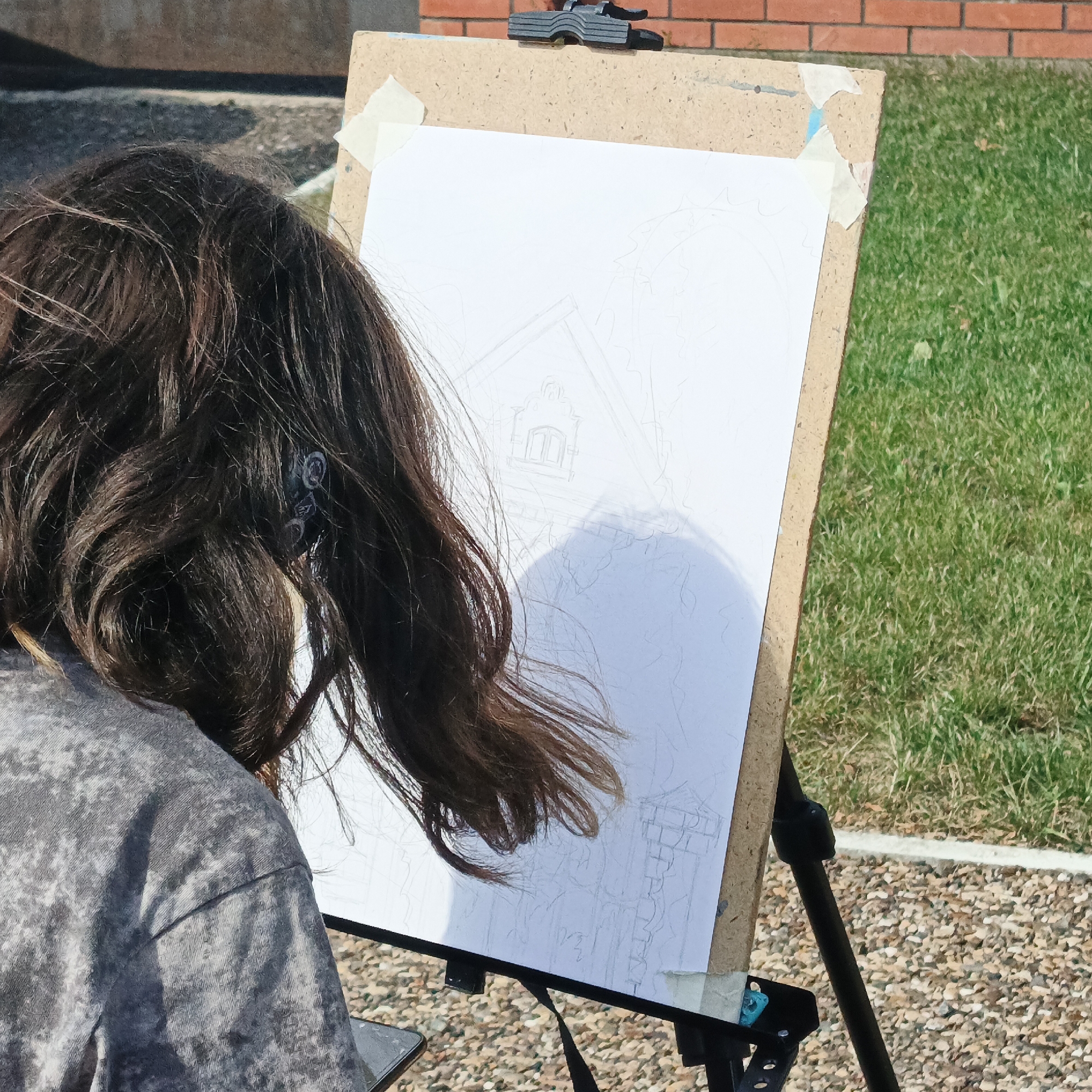 An over-the-shoulder medium shot captures a person, whose face is obscured by their dark, wavy hair, working on a sketch of a building at an easel outdoors. The person is wearing a grey t-shirt. The easel is positioned on a gravel surface, with green grass and a brick wall visible in the background. The sketch on the paper depicts a building with a window and architectural details, and a shadow is cast across the paper. The scene appears to be in an outdoor setting, possibly a park or yard, during daylight. The overall impression is one of quiet focus and creative activity.