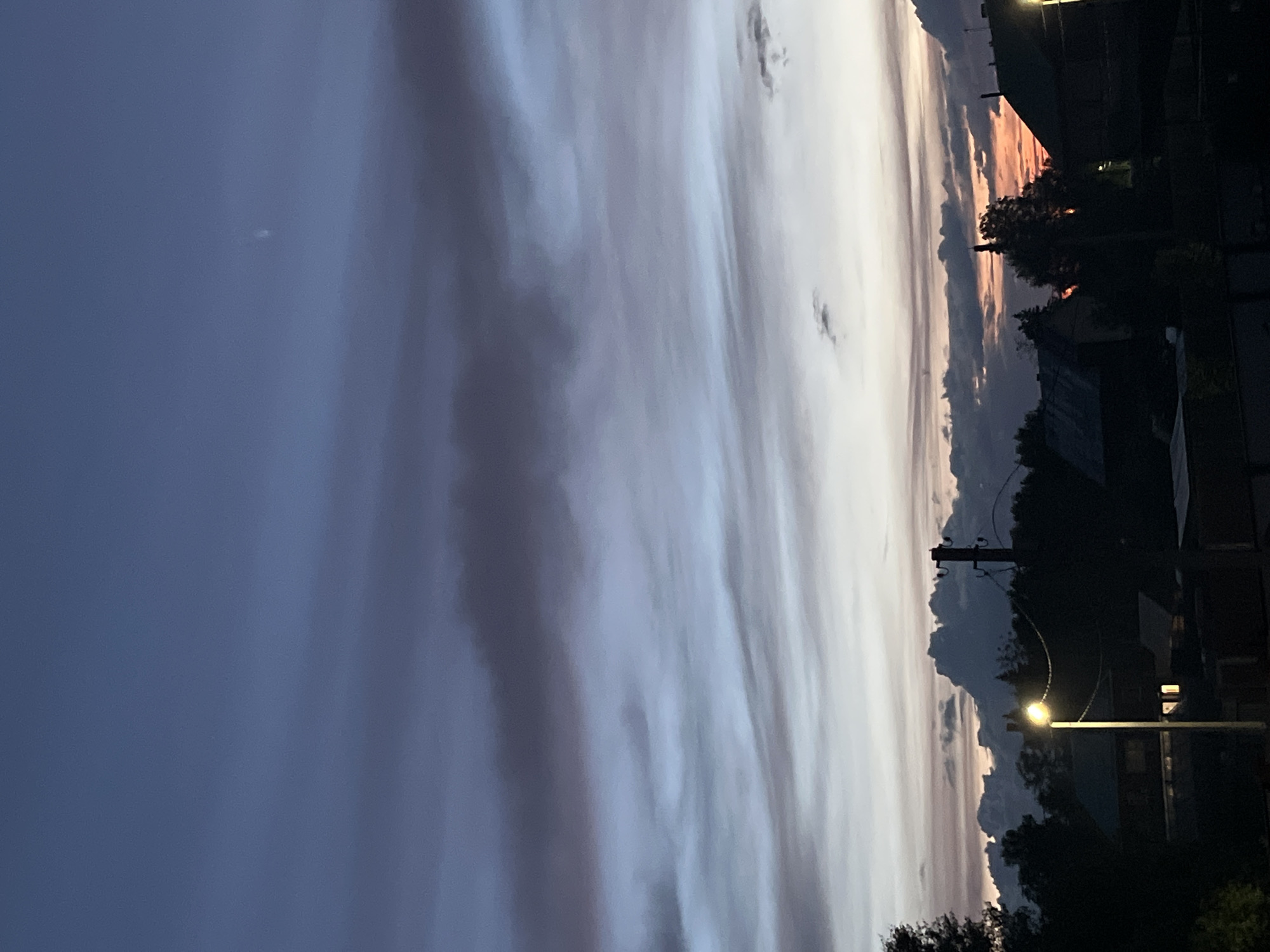 The image captures a dusk or dawn scene in Chaikovsky, Russia. The sky is filled with elongated, striated clouds displaying a range of colors from deep blue to pale white with hints of pink and orange near the horizon. The foreground consists of silhouetted buildings, trees, and utility poles, indicating an urban or suburban setting. A street light is illuminated, casting a warm glow on its immediate surroundings.