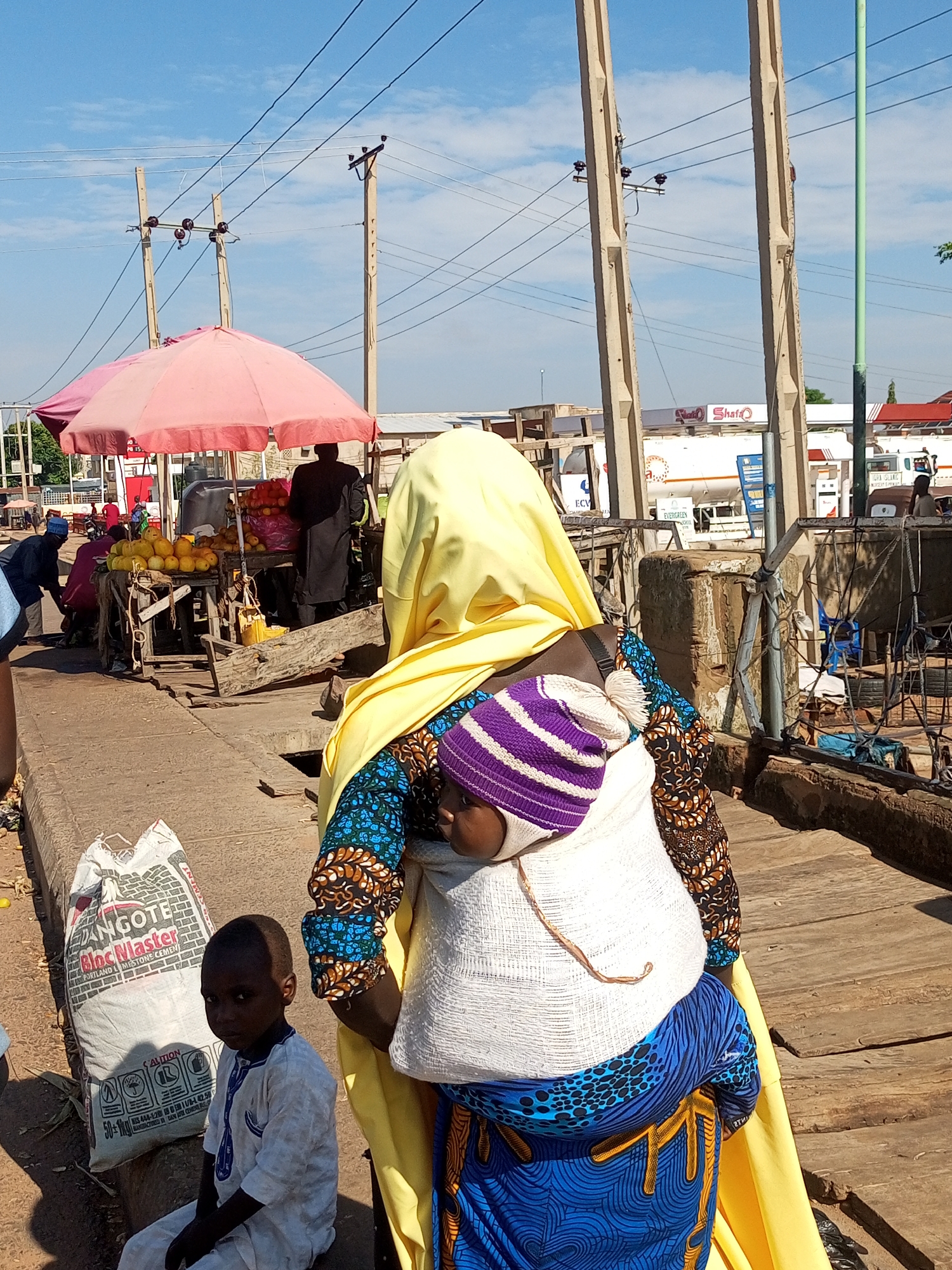 In an outdoor market scene in an unknown city, a woman wearing a yellow headscarf and a colorful patterned dress is pictured from the back, carrying a baby on her back. The baby is wearing a striped hat. Next to them, a young boy in a white outfit is sitting near a large white bag. In the background, a fruit stall with pink umbrellas is visible, along with people and other market activity. Power lines and poles are prominent in the sky above.