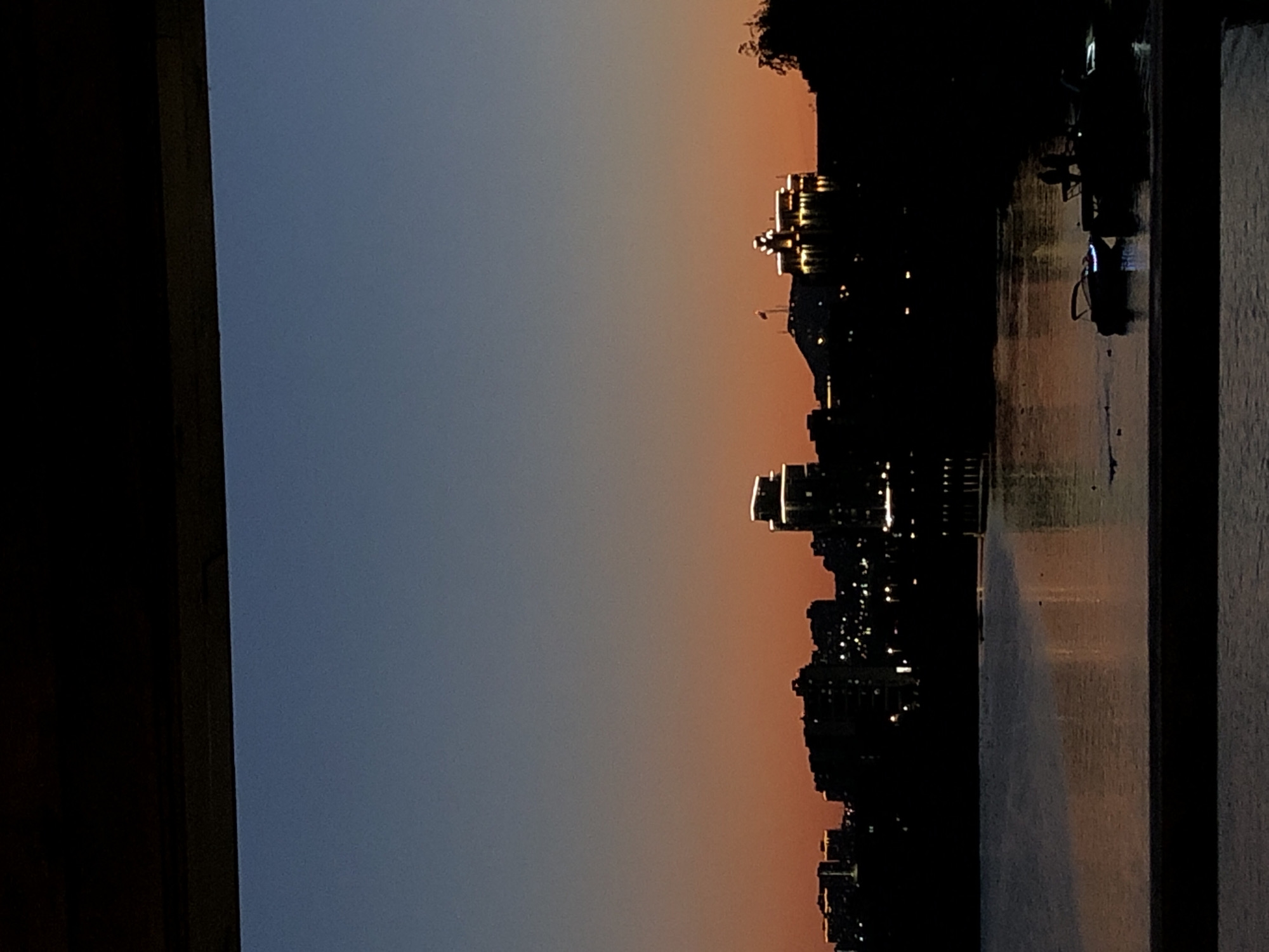The image depicts a twilight cityscape with a body of water in the foreground and a silhouetted skyline in the background. A dark, horizontal structure, possibly a railing or balcony, frames the bottom of the scene, and a similar dark structure is visible at the top, obscuring the sky. The sky transitions from a deep blue at the top to an orange hue near the horizon.
The skyline features several buildings, some of which are illuminated, creating a warm glow against the darkening sky. One prominent building in the mid-right of the skyline is lit with distinct architectural lighting. Several smaller buildings with scattered lights are visible to the left.
On the water's surface, reflections of the city lights are visible. Several boats are present on the water. In the mid-right of the water, a boat with a blue light is moored next to a dock. Two figures are visible on the dock, silhouetted against the water. Other smaller boats and possible ducks are also present on the water. The overall impression is of a serene evening scene in a city.