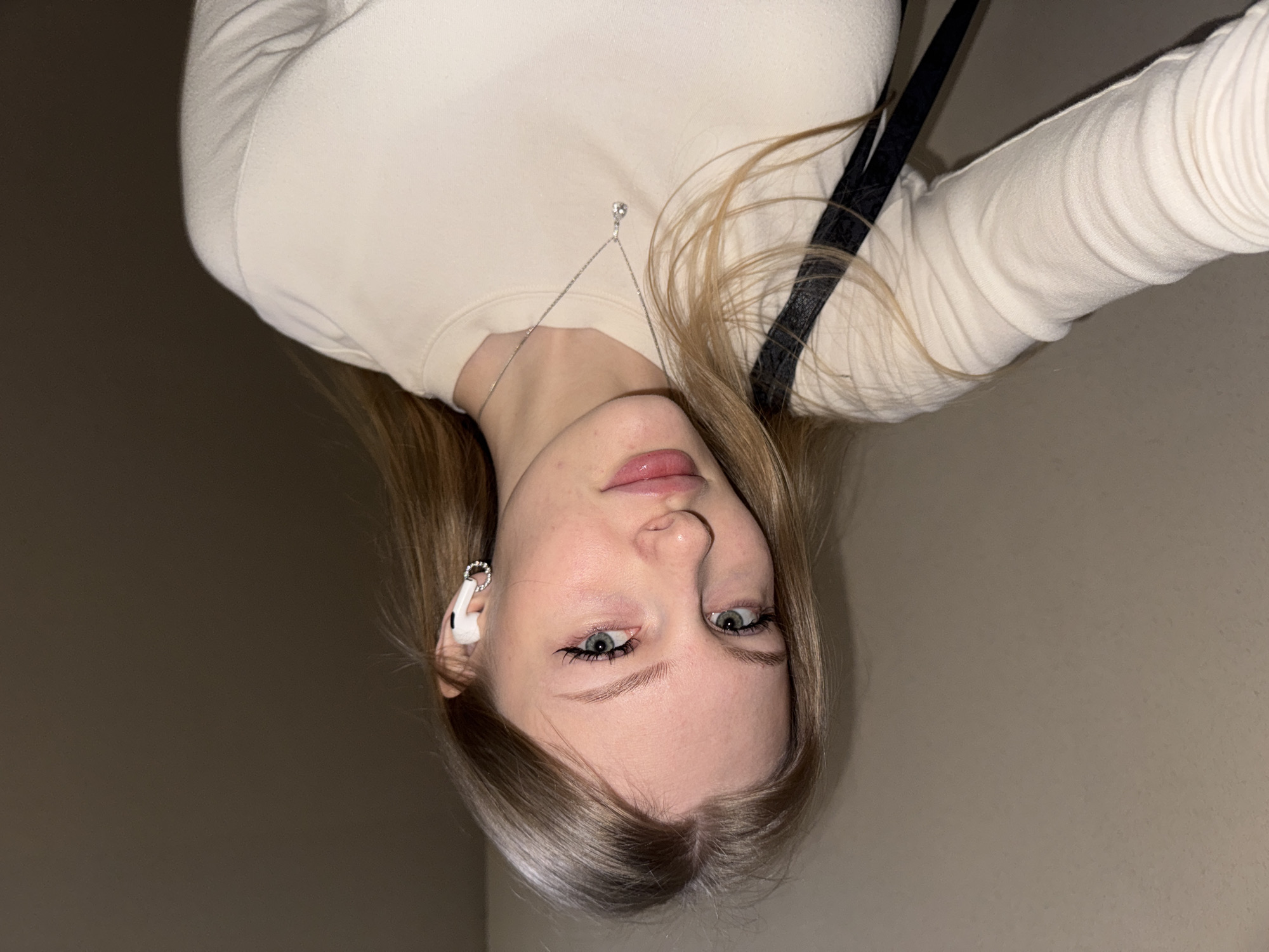 A young, fair-skinned woman with blonde hair and green eyes poses for a selfie. She is wearing a light beige long-sleeved shirt and a silver necklace with a pendant. A black strap is visible on her shoulder. She has a white wireless earbud in her left ear. The background is a plain beige wall. There is no discernible information to indicate the location is Perm, Russia.