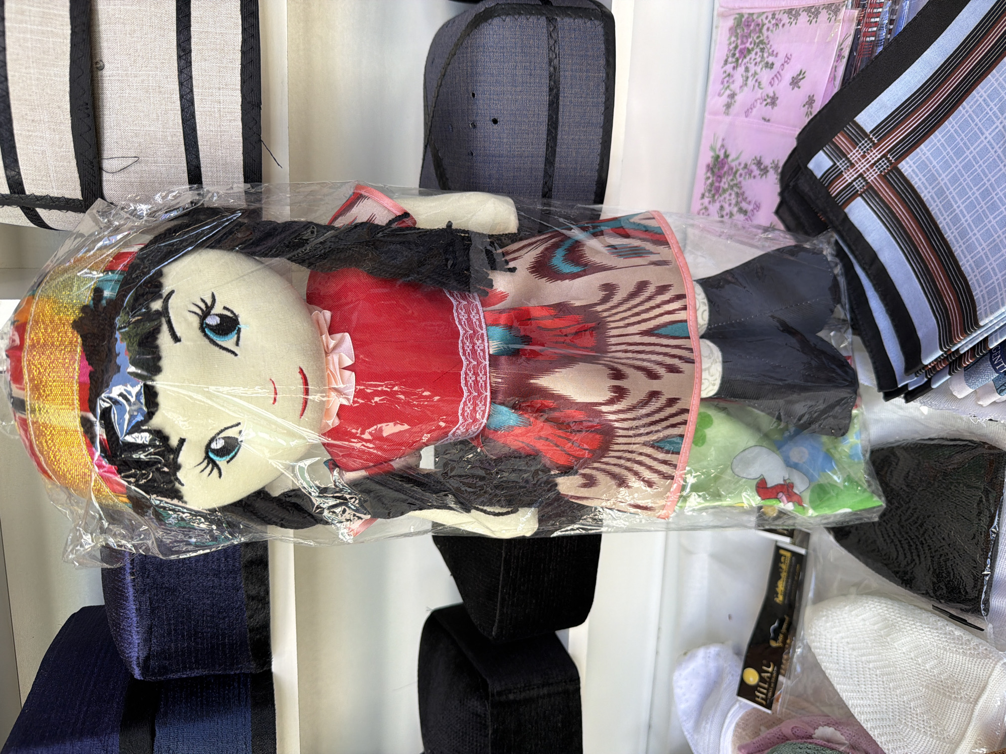 A plush doll, wearing a red top and a patterned skirt, is displayed in plastic packaging in the foreground. The doll has black braided hair, blue eyes, and a red mouth. It stands in front of shelves filled with various caps. On the left, several dark blue caps with black trim are visible. To the right of the doll, several black and blue caps are also present. In the background, more caps with black trim are stacked. At the bottom of the image, to the left of the doll, are white and black knit caps, along with a packaged item labeled "Hilal". To the right, several folded fabric squares, some with floral patterns and others with plaid designs, are stacked. The overall setting appears to be a retail environment, likely a market stall or shop. The presence of the caps, some of which appear to be kufi or similar religious headwear, suggests a cultural context. The doll's attire, particularly the patterned skirt and colorful headpiece, might indicate traditional Uzbek clothing. The scene is lit by overhead lighting, casting soft shadows. The image is taken at a slight upward angle, emphasizing the display of goods. This image was captured in the city of Zangiata, Uzbekistan.