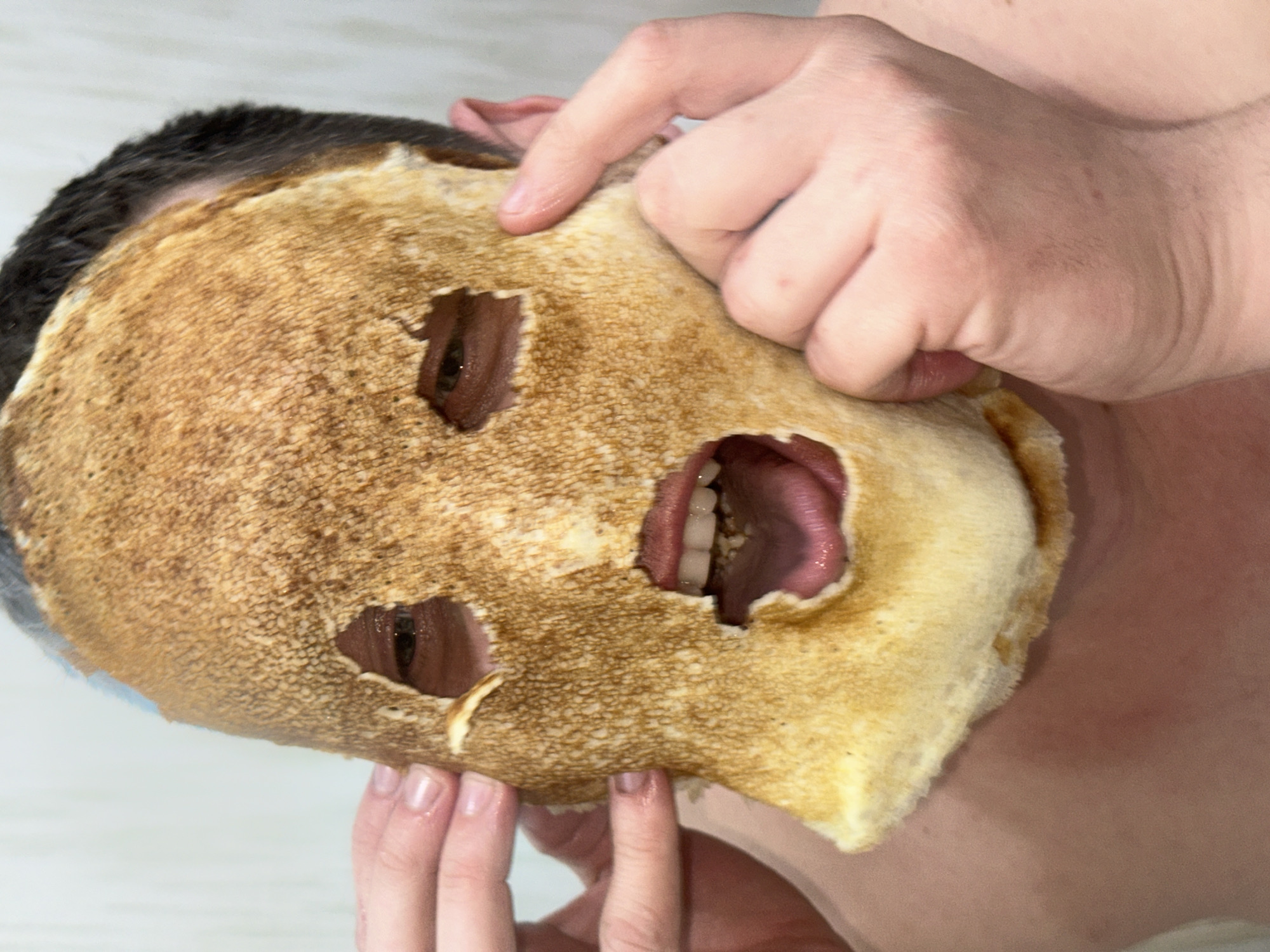 A person is holding a pancake over their face. The pancake has holes cut out for the eyes and mouth. The person's eyes are visible through the eye holes and their tongue is visible through the mouth hole. The person's hair is dark. The environment is a room with white walls. There is no indication as to the location within the city of Chaikovsky, Russia.