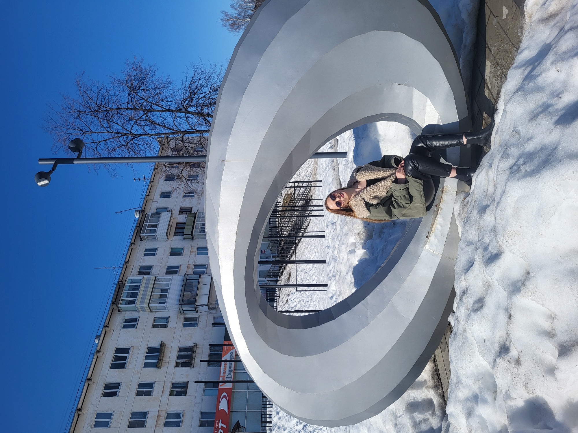 A woman is sitting inside a large, circular, silver sculpture in the city of Chaikovsky, Russia. The sculpture is comprised of several concentric rings. The woman is wearing black boots, black pants, and a green jacket with a fuzzy, light-colored collar. The sculpture is situated on a patch of snow. Behind the sculpture is a multi-story, white building with numerous windows and balconies. A bare tree is visible beside the building. A street lamp stands to the left of the building. The sky is clear and blue.