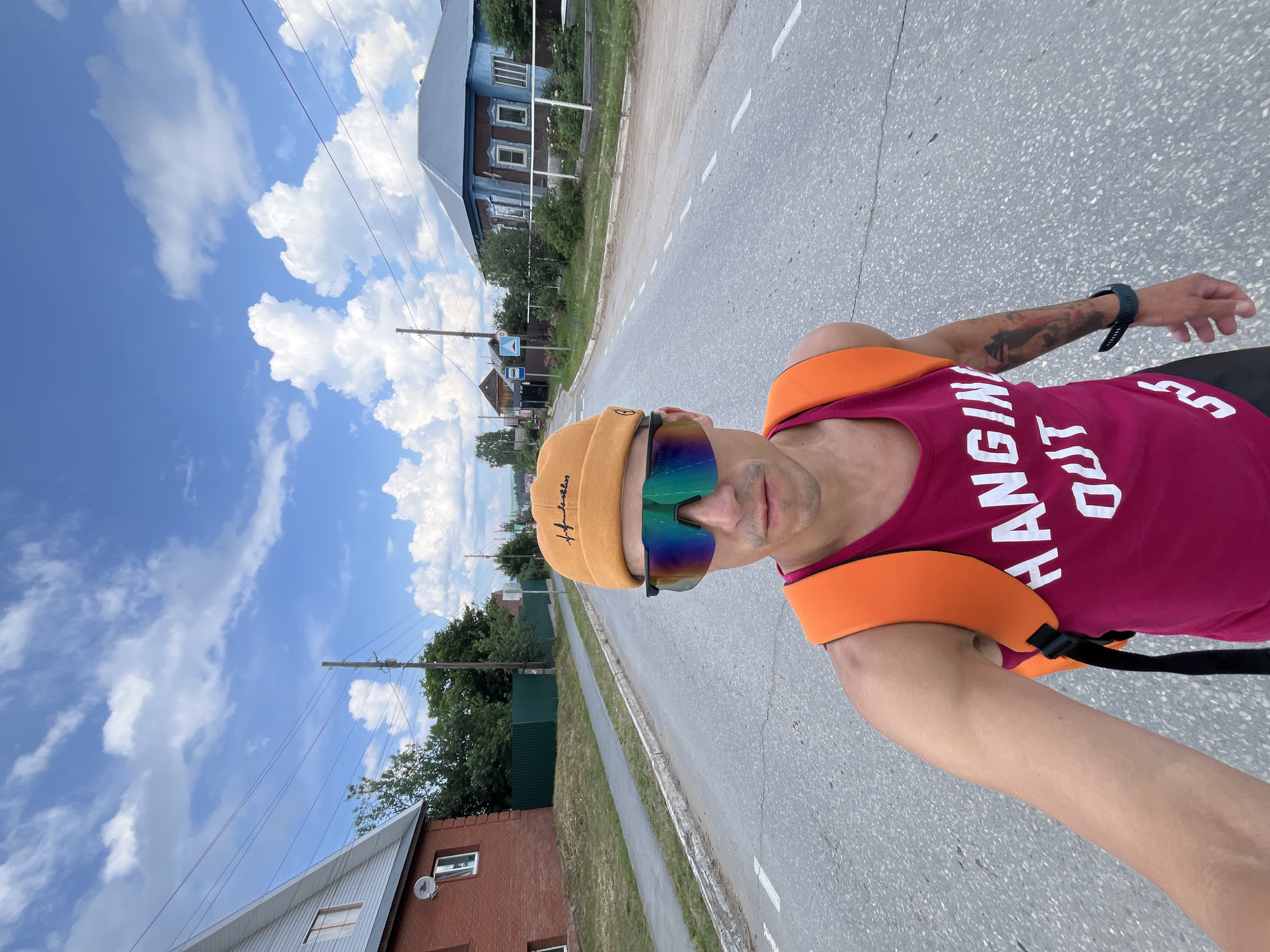 A person wearing a pink tank top, orange backpack, and orange beanie takes a selfie on a paved road in Chaikovsky, Russia. The person is also wearing multicolored sunglasses and a dark smartwatch. The road is lined with houses and trees, and power lines are visible overhead. The sky is blue with white clouds.