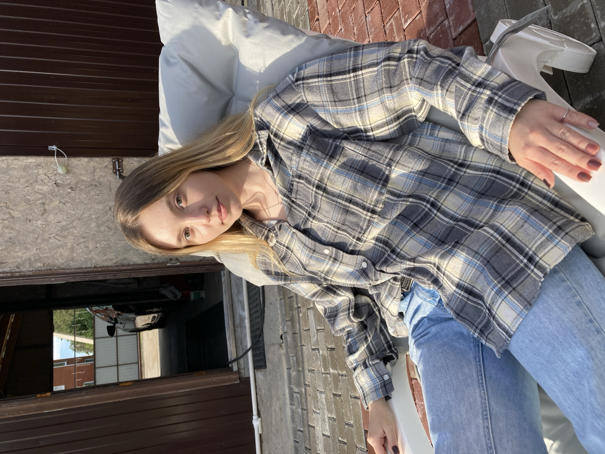 A woman with light brown hair is reclining in a white chair outdoors. She is wearing a gray, blue, and white plaid shirt and light blue jeans. A garage door is visible behind her to the left. The location is Chaikovsky, Russia.
