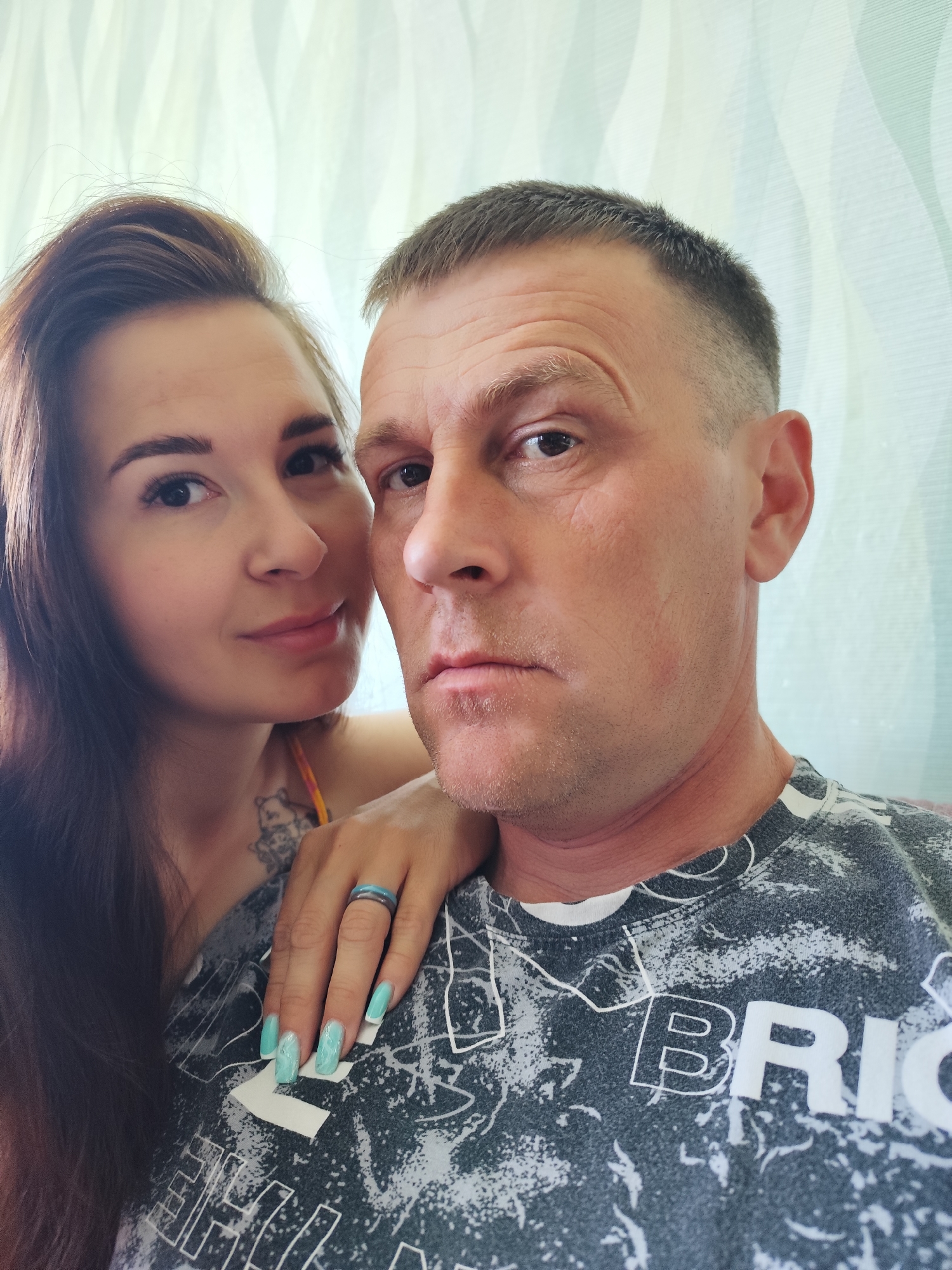 A selfie shot features a woman and a man posing closely together. The woman, with long, dark hair, stands to the left and has a tattoo on her chest. She rests her hand on the man's shoulder; the hand features long, painted nails and a ring. The man has a short haircut and is wearing a patterned T-shirt with the word "BRICK" visible. The background appears to be an interior setting with textured wallpaper. There is no explicit indication of the city Фоки, Russia in the image.