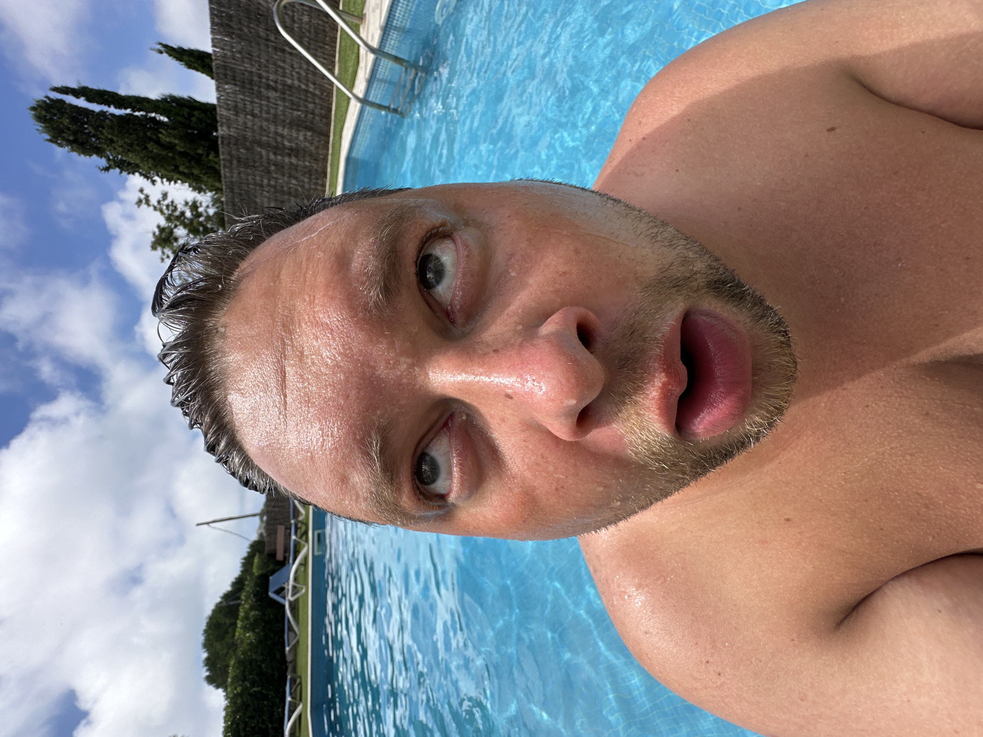 The image shows a close-up, fisheye perspective of a man's face partially submerged in a bright blue swimming pool. The man's hair is wet and slicked back, and his face is glistening with water droplets. His eyes are rolled upwards, and his mouth is slightly open, giving him a surprised or bewildered expression. The background features the pool's edge, a tiled bottom, a metal ladder, and lush green foliage and trees beyond a fence. The sky above is a bright blue with scattered white clouds. The scene appears to be outdoors on a sunny day, likely in a residential or resort setting within Sant Feliu de Guíxols, Spain.