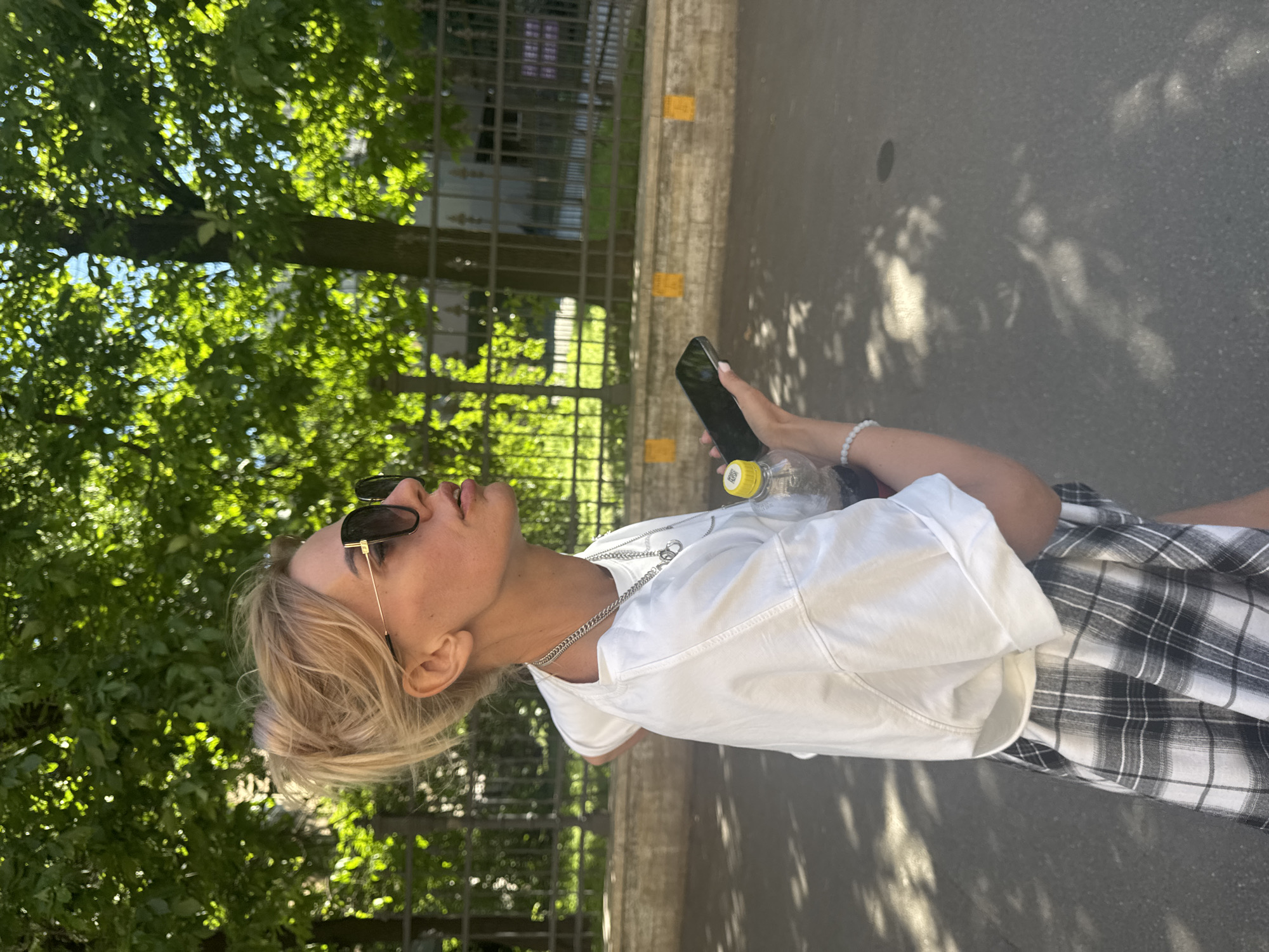 A woman with blonde hair is standing outdoors, holding a smartphone in her right hand and a water bottle in her left. She is wearing sunglasses, a white t-shirt, and a plaid skirt. The background features lush green trees, a metal fence, and a paved path with dappled sunlight and shadows. The scene appears to be set during the day, likely on a sunny day. The woman's gaze is directed upwards, suggesting she might be looking at something above or lost in thought. There are no discernible text elements in the image.